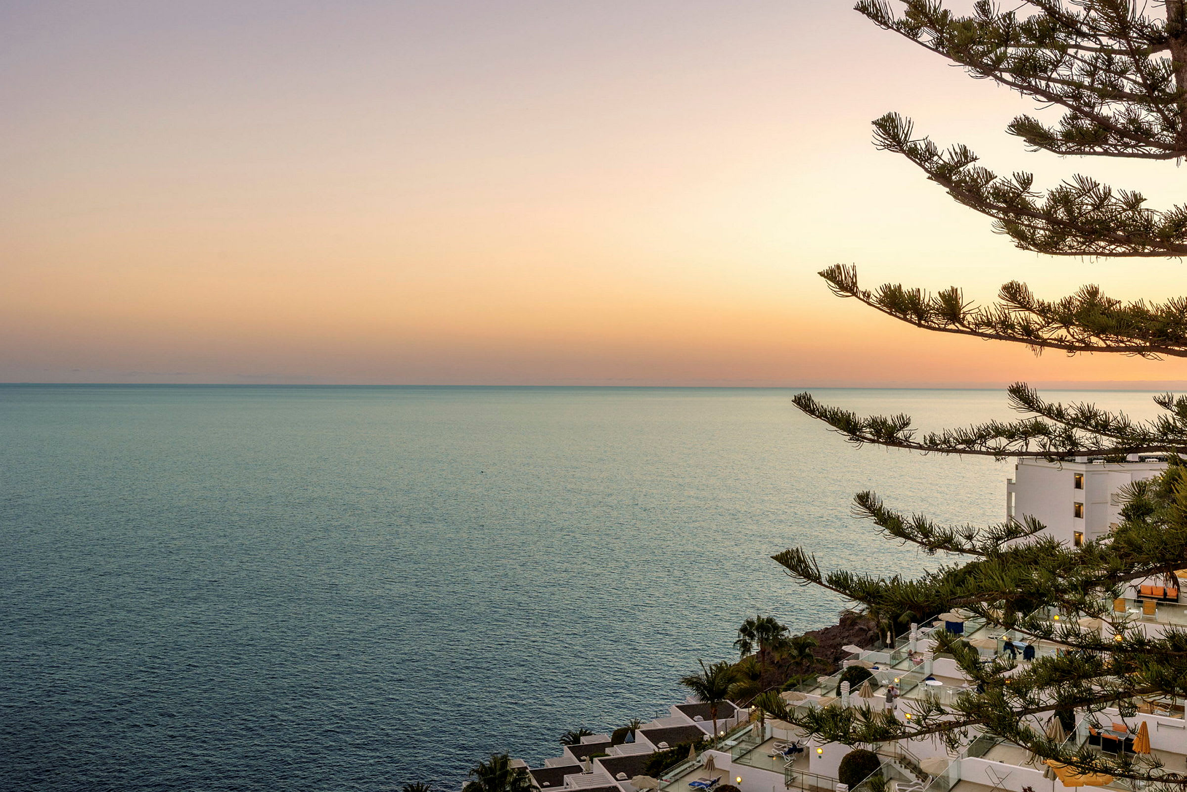Solnedgang over havet med en bygning og trær i forgrunnen. (AI-generert tekst)