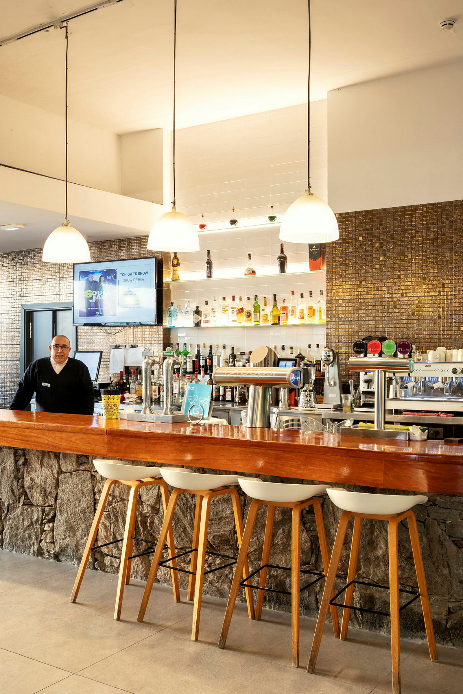 En hotellbar med trekantdisk og stoler, samt hyller fylt med flasker. (AI-generert tekst)
