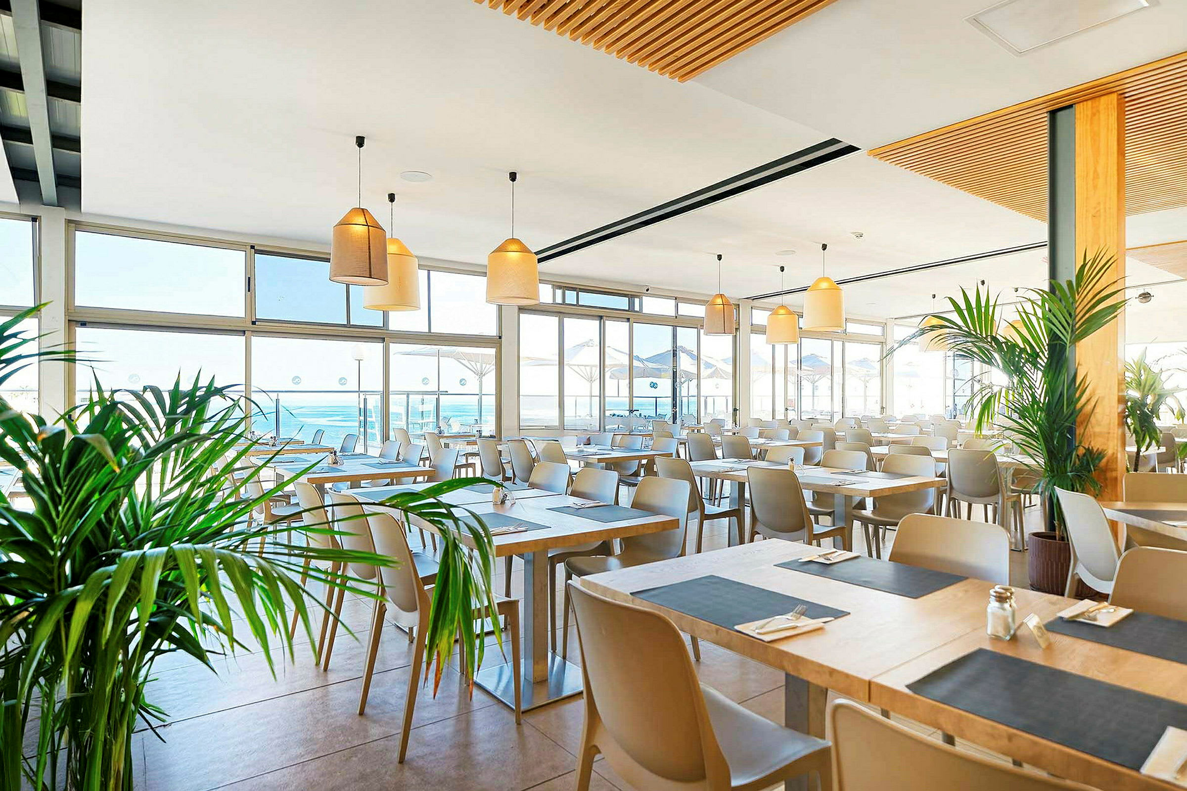 Lys og moderne restaurant med mange bord, store vinduer og planter. (AI-generert tekst)