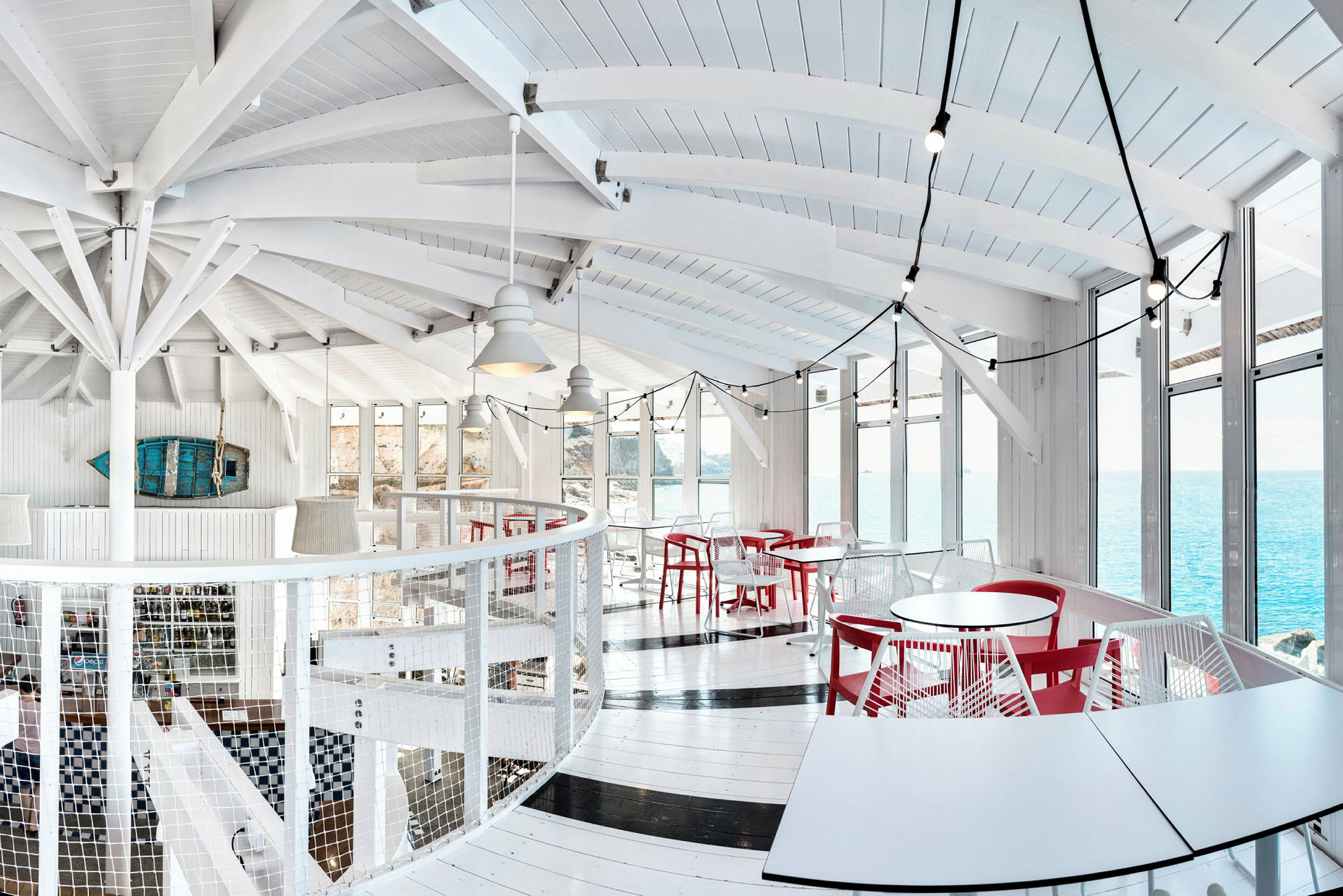 Lyst innredet restaurant med hvite vegger, røde stoler og store vinduer med utsikt over havet. (AI-generert tekst)