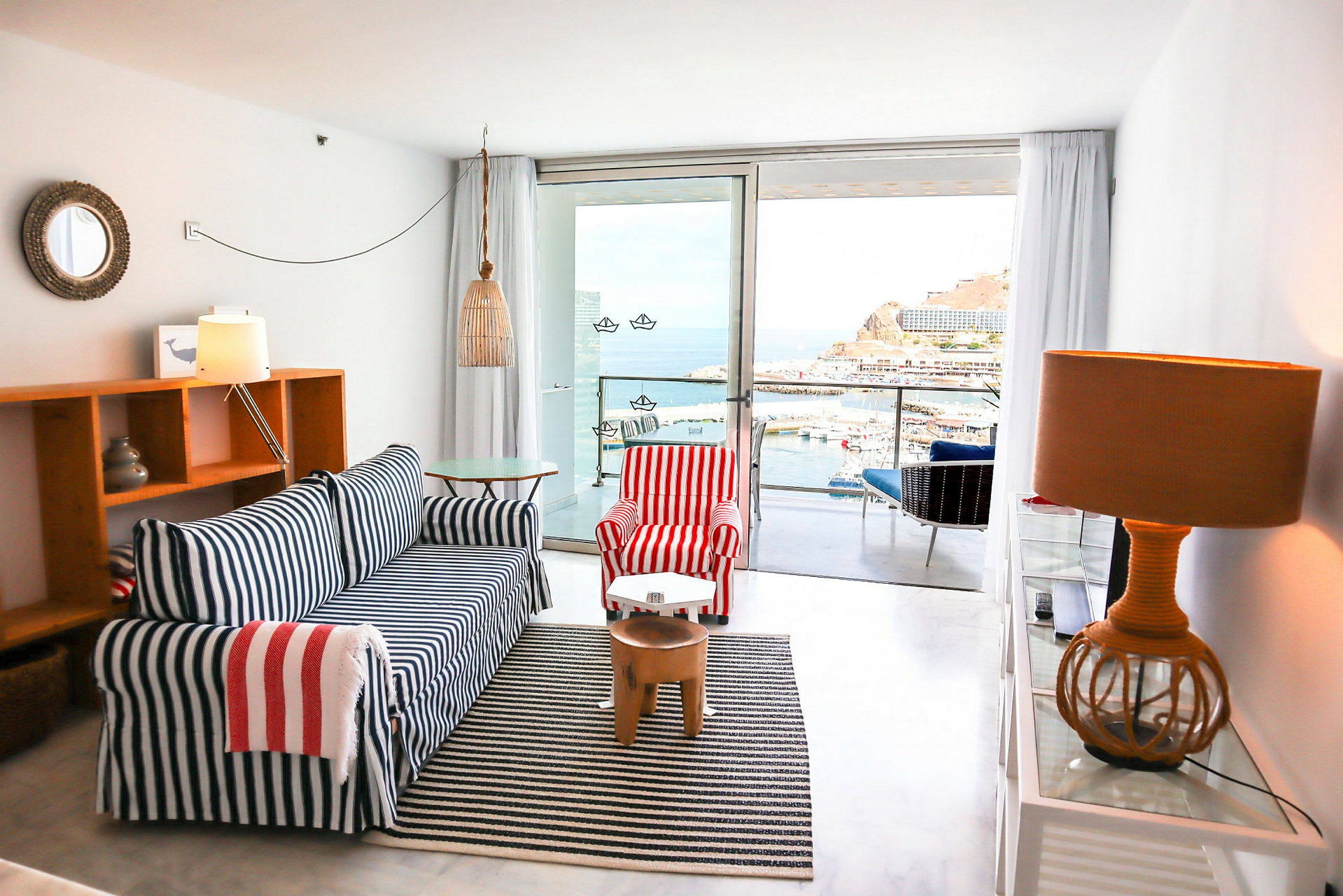 Et hotellrom med maritim innredning, sofaen har blå og hvite striper, lenestolen har røde og hvite striper. Utsikt mot en havn og hav fra en balkong. (AI-generert tekst)