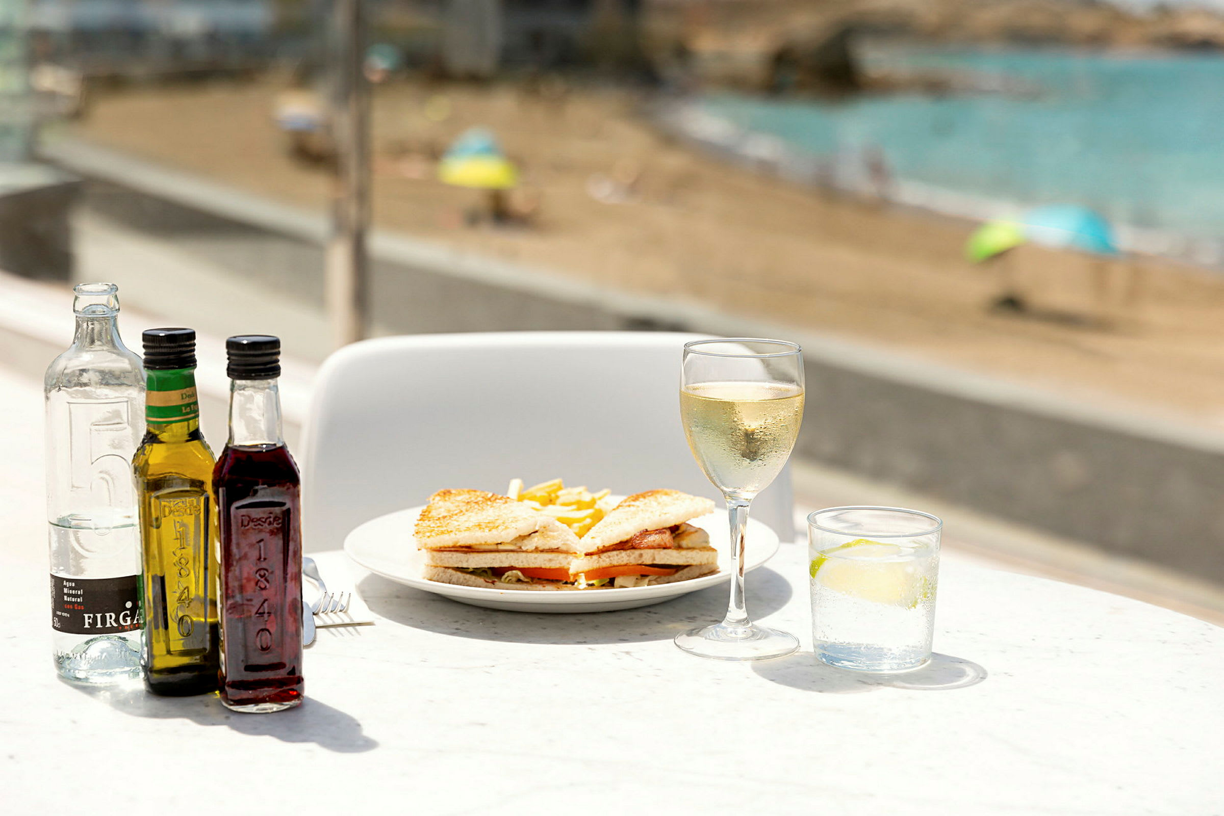En tallerken med sandwich og pommes frites, omgitt av flasker, et glass hvitvin og et glass vann med lime, med en strand i bakgrunnen. (AI-generert tekst)