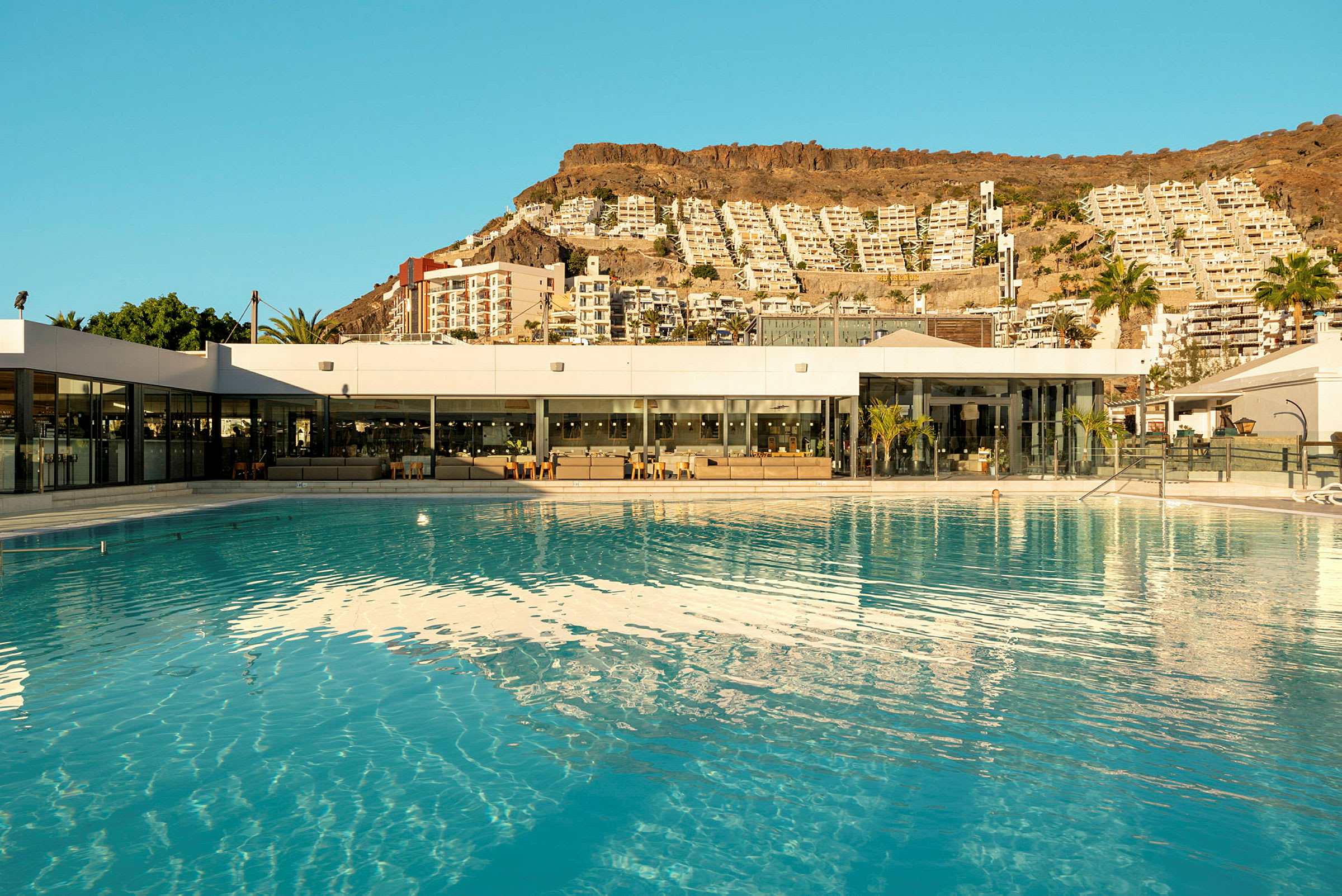 Stor utendørsbasseng ved moderne bygning, omgitt av landskap med høyhus på en fjellskråning. (AI-generert tekst)