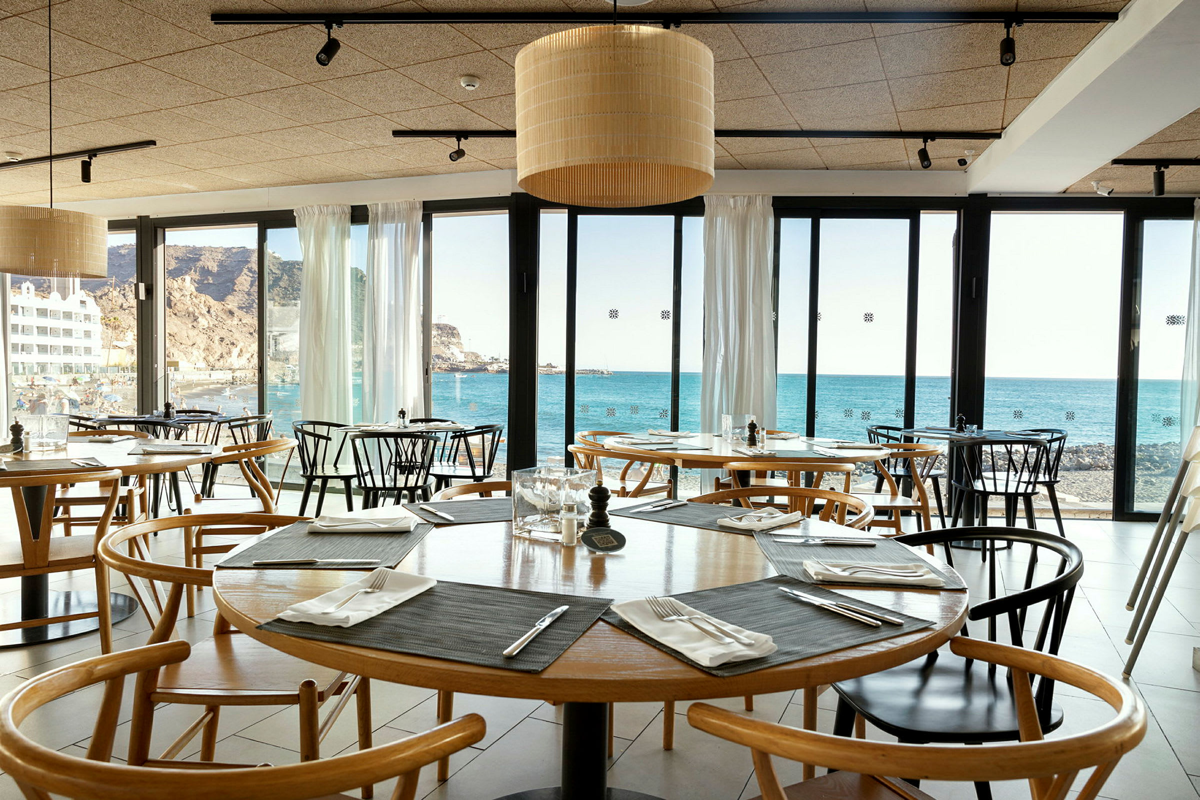 Innsiden av en restaurant med bord og stoler, utsikt mot havet gjennom glassvinduer. (AI-generert tekst)