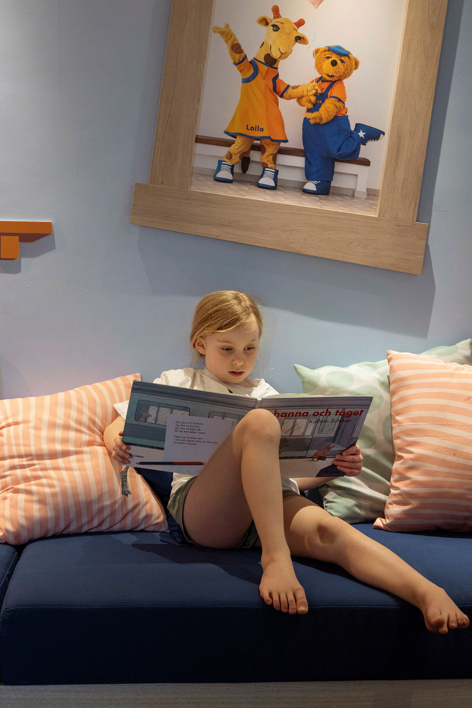 Et barn sitter på en sofa og leser en bok, med et maleri som viser dyrefigurer i bakgrunnen. (AI-generert tekst)