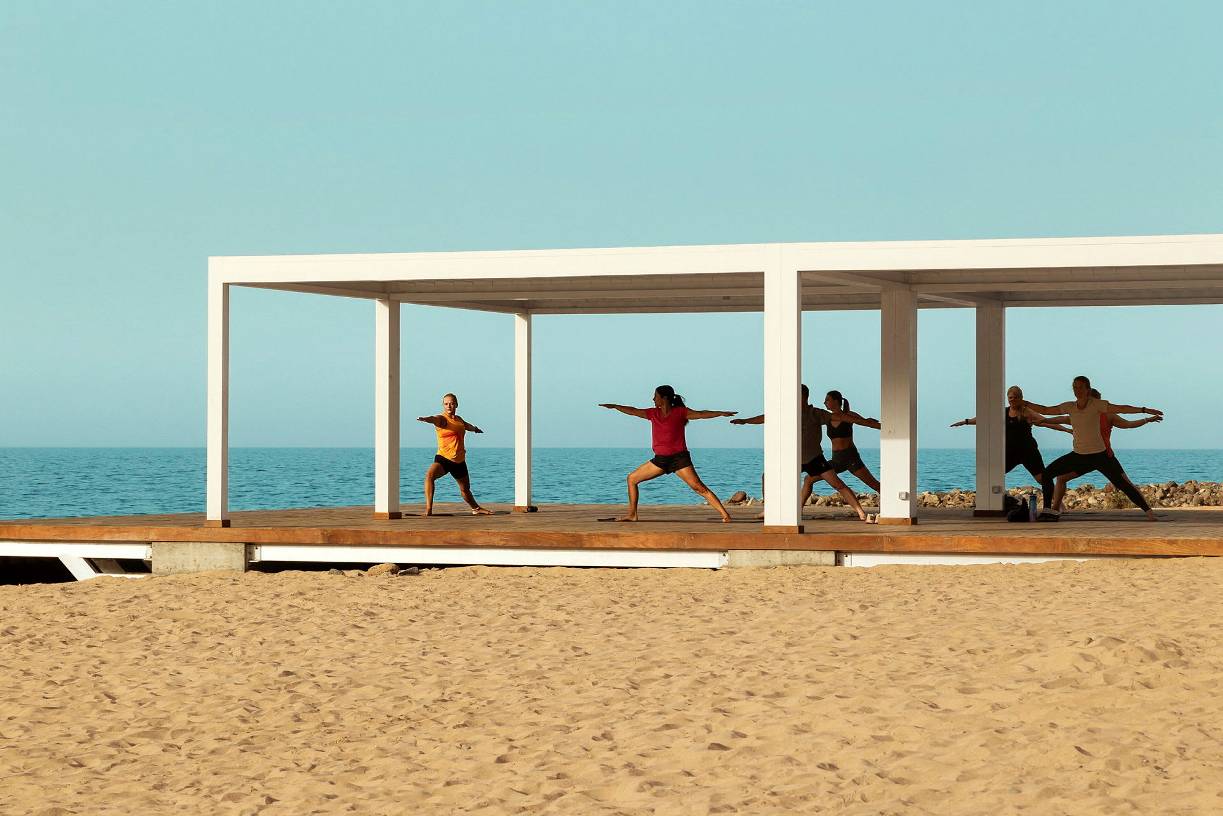 Personer som utfører yoga på en treplattform ved havet under en hvit pergola. (AI-generert tekst)
