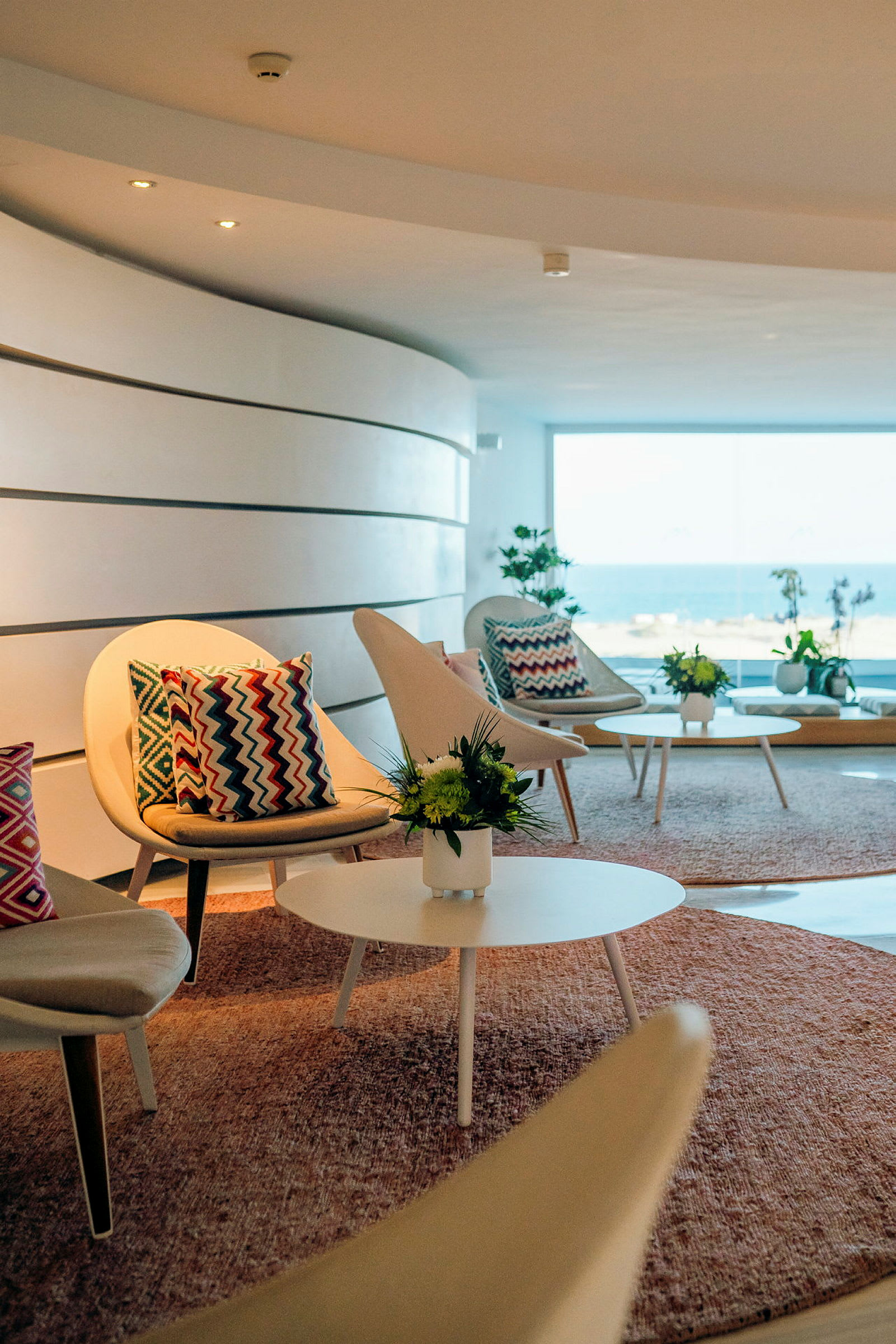 En lounge med moderne stoler og sofabord, dekorert med fargerike puter og blomster, med havutsikt gjennom et stort vindu. (AI-generert tekst)