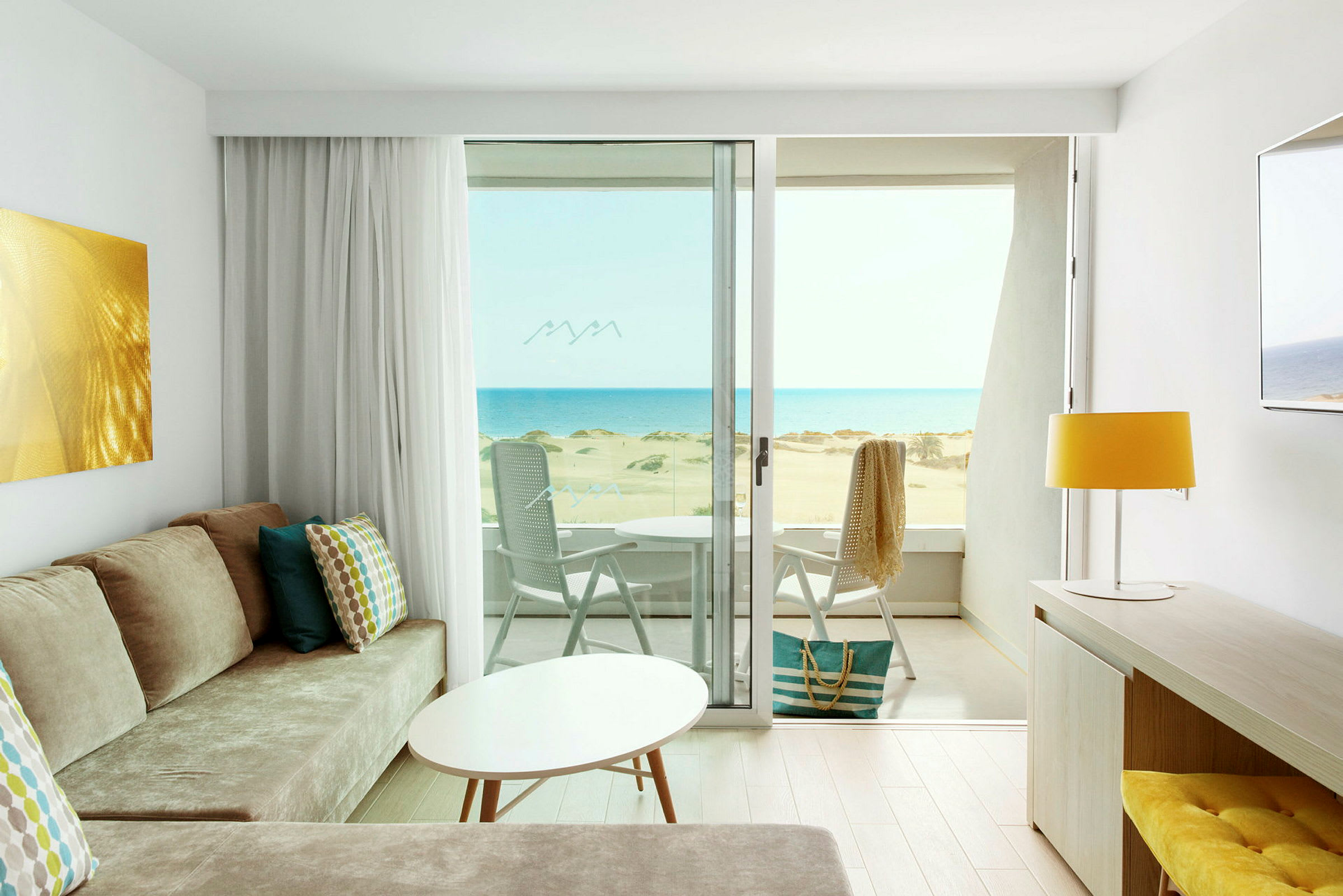 Utsikt fra et lyst hotellrom med moderne innredning over en solfylt strand og havet. (AI-generert tekst)