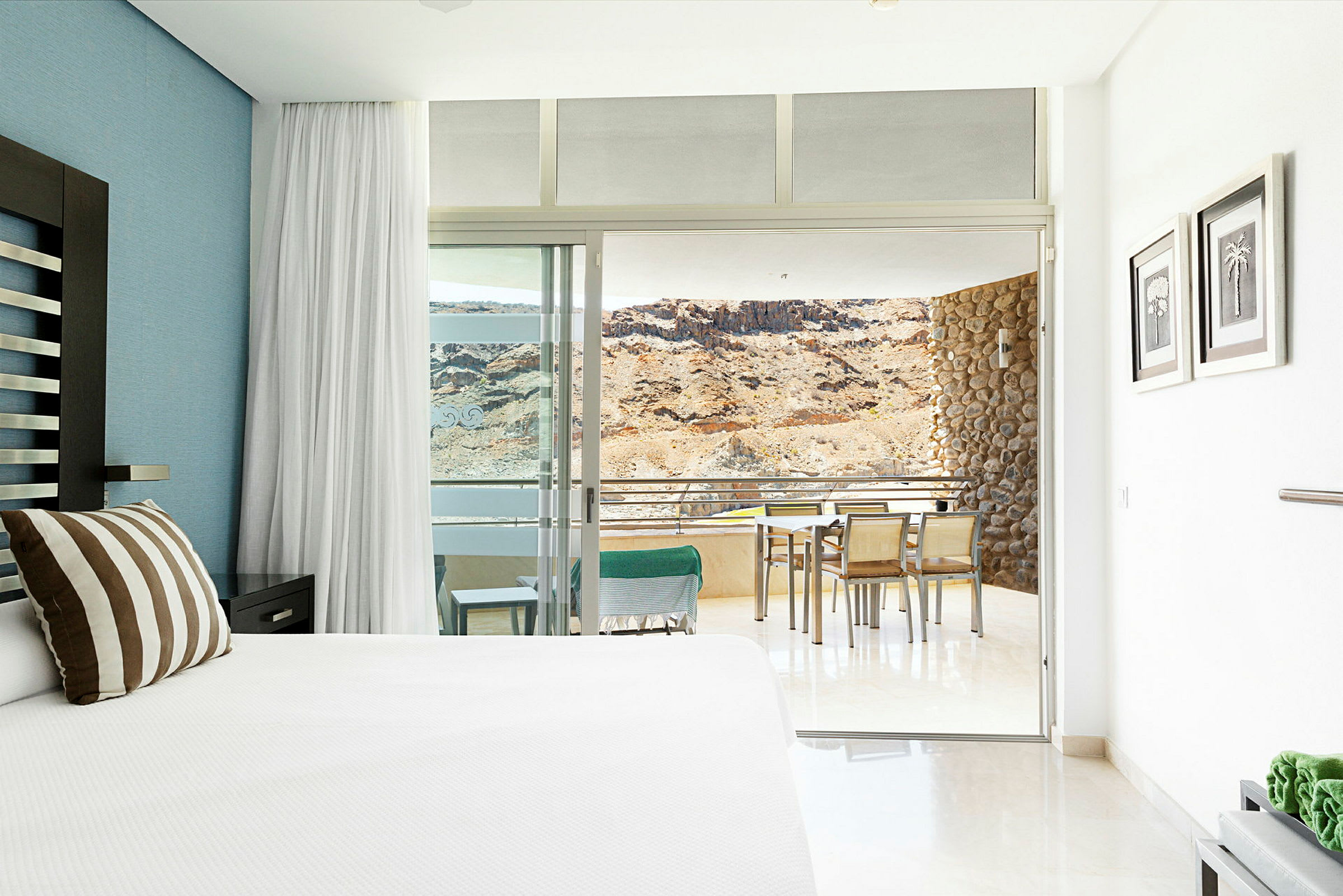 Hotellrom med en stor seng og utsikt mot en terrasse med bord og stoler samt en bakgrunn med fjell. (AI-generert tekst)