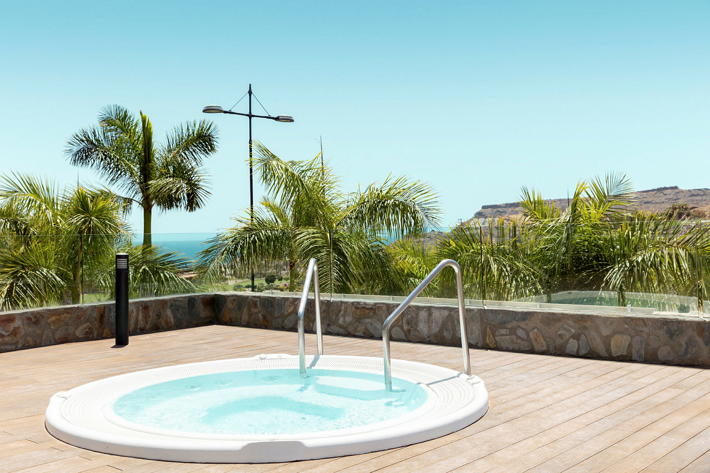 Rund jacuzzi på en terrasse med havutsikt og palmer. (AI-generert tekst)