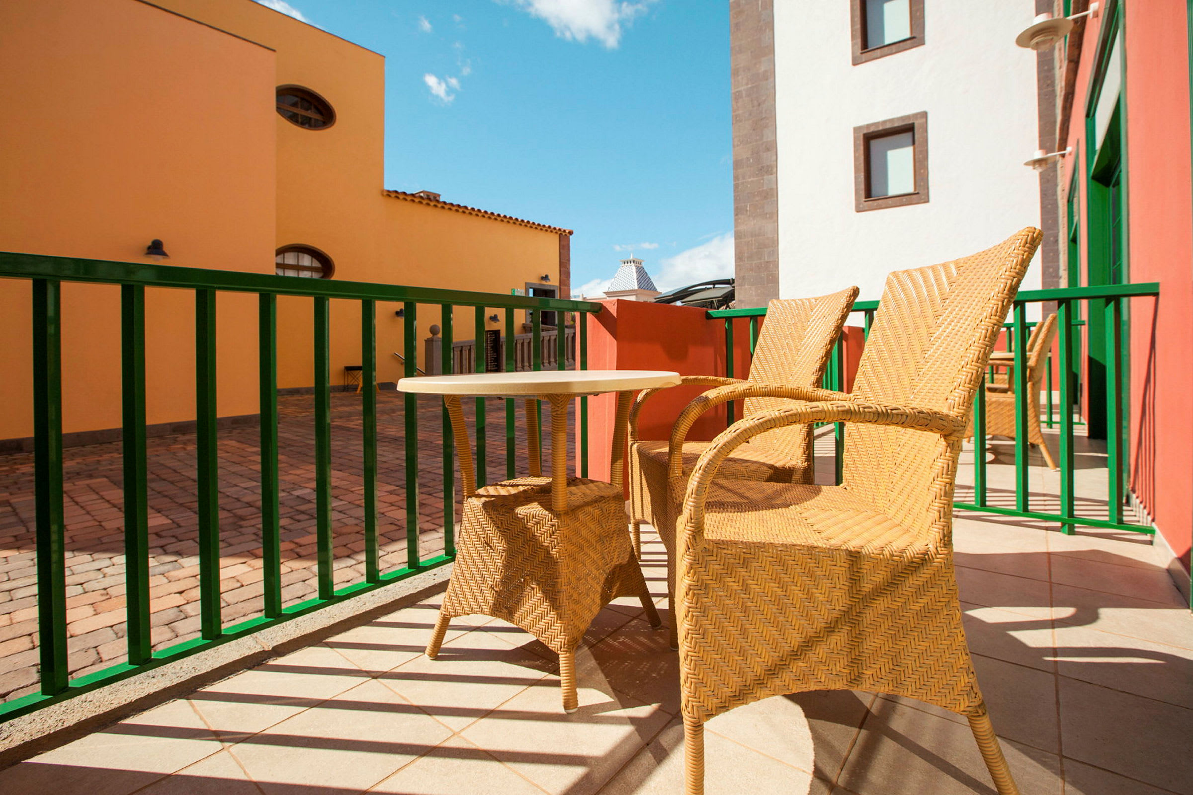 To flettede stoler og et lite bord står på en solfylt terrasse med fargerike bygninger i bakgrunnen. (AI-generert tekst)