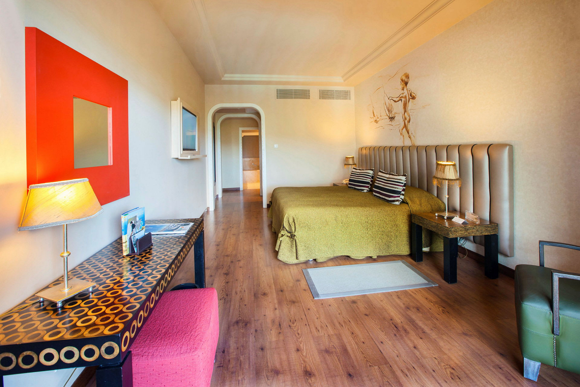 Et hotellrom med tremønstrede gulv, en grønn oppredt seng, et skrivebord med lampe og dekorasjoner, samt en veggmontert TV. (AI-generert tekst)