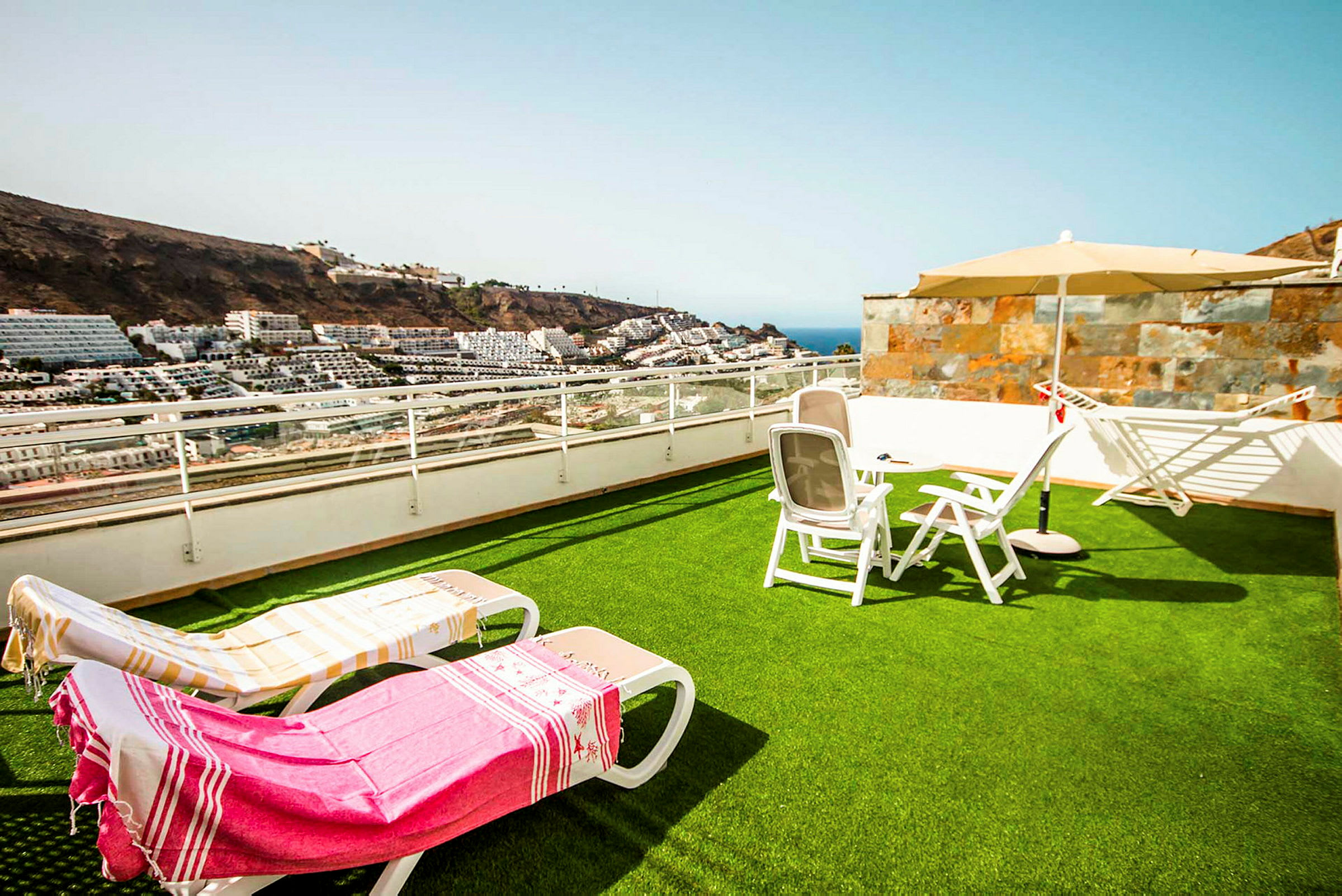 En terrasse med gressplen, solsenger og parasoll, med utsikt over havet og omkringliggende bygninger. (AI-generert tekst)
