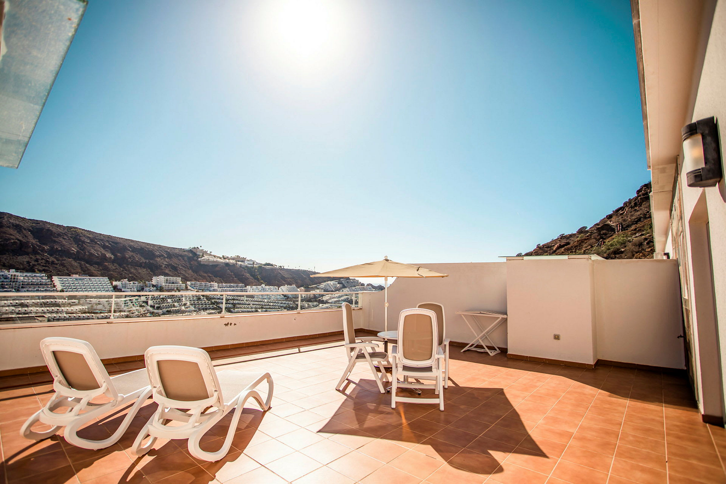 Solfylt terrasse med utemøbler og utsikt mot omkringliggende bygninger og fjell. (AI-generert tekst)