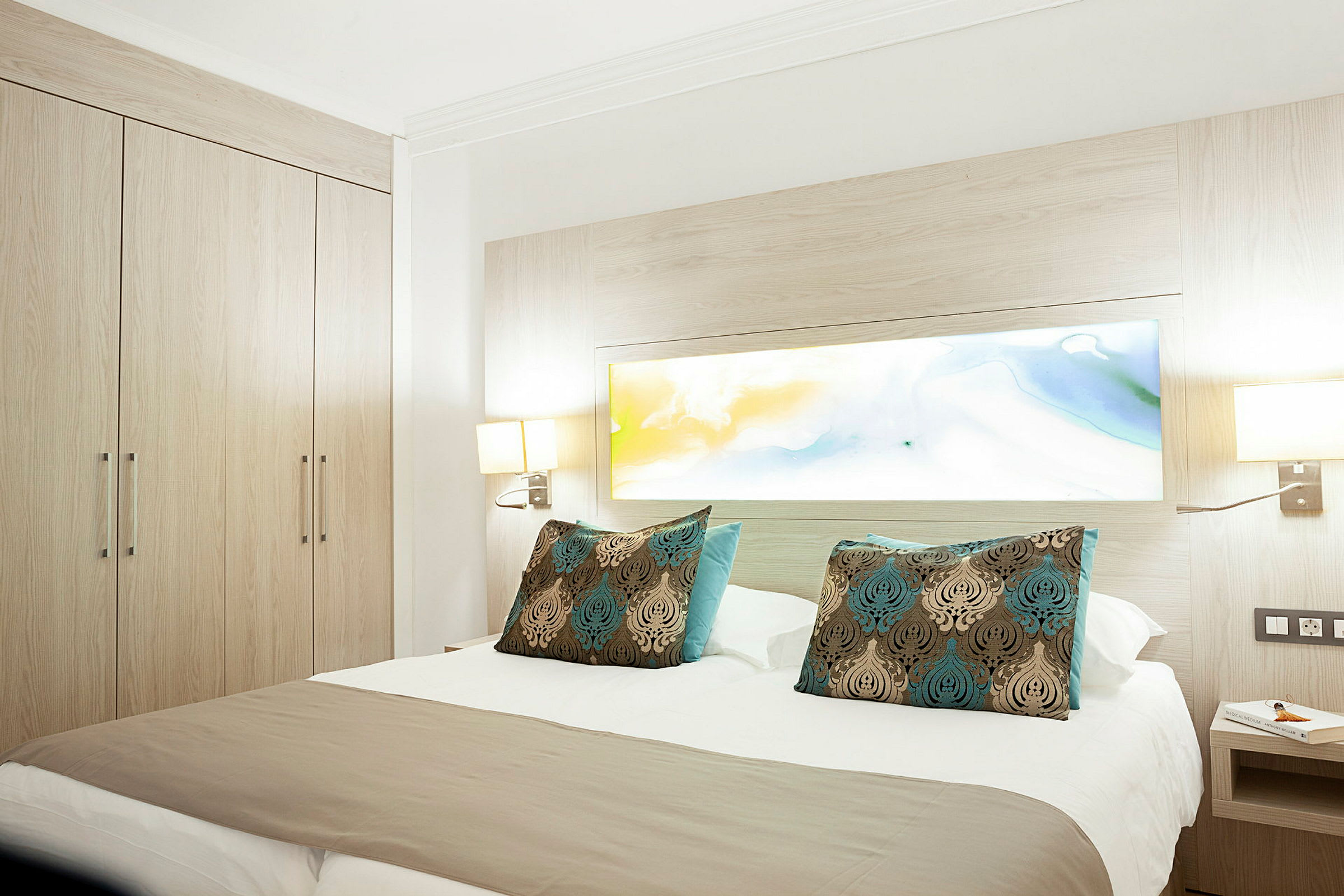 Hotellrom med en dobbeltseng og dekorative puter, omringet av lyst treverk og lamper. (AI-generert tekst)
