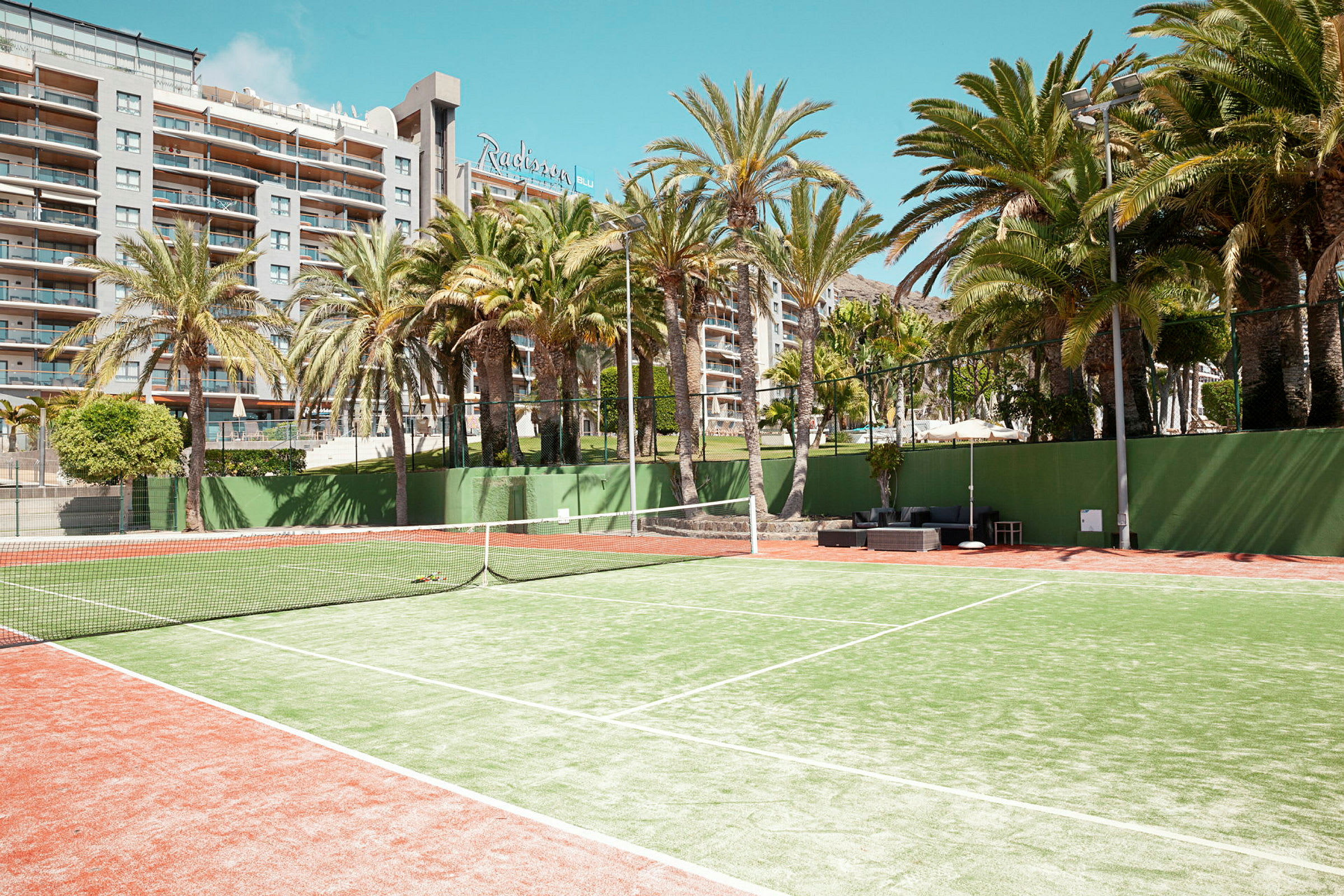 Tennisbane med grusunderlag omgitt av palmer, med et hotell i bakgrunnen. (AI-generert tekst)