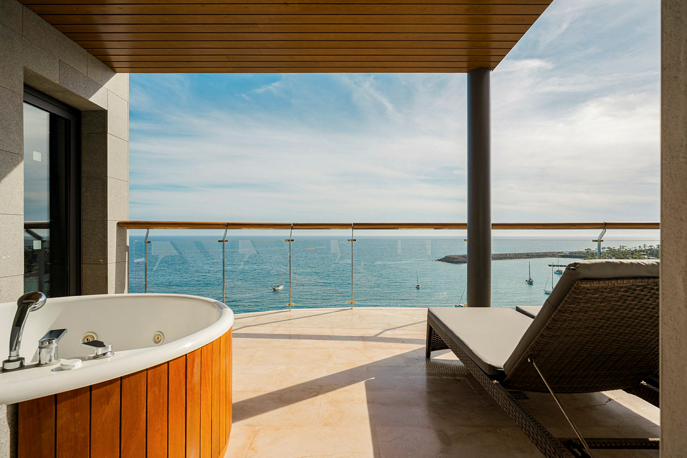En hotellbalkong med boblebad og solseng, utsikt over havet og småbåter. (AI-generert tekst)