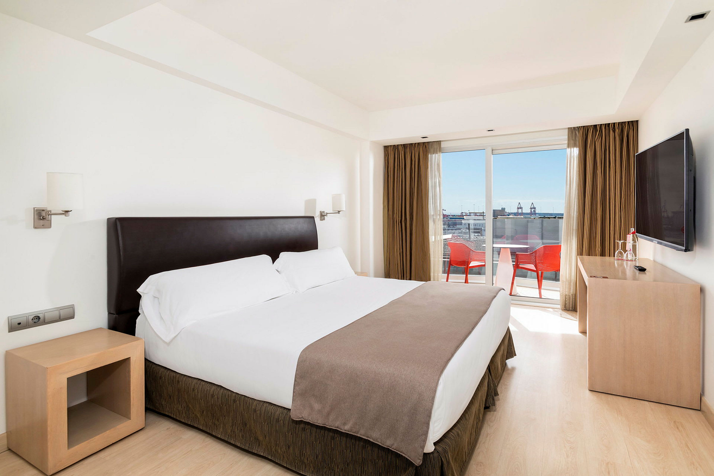 Hotellrom med en dobbeltseng, nattbord og TV. Vindu med utsikt mot en balkong med stoler og bord. (AI-generert tekst)