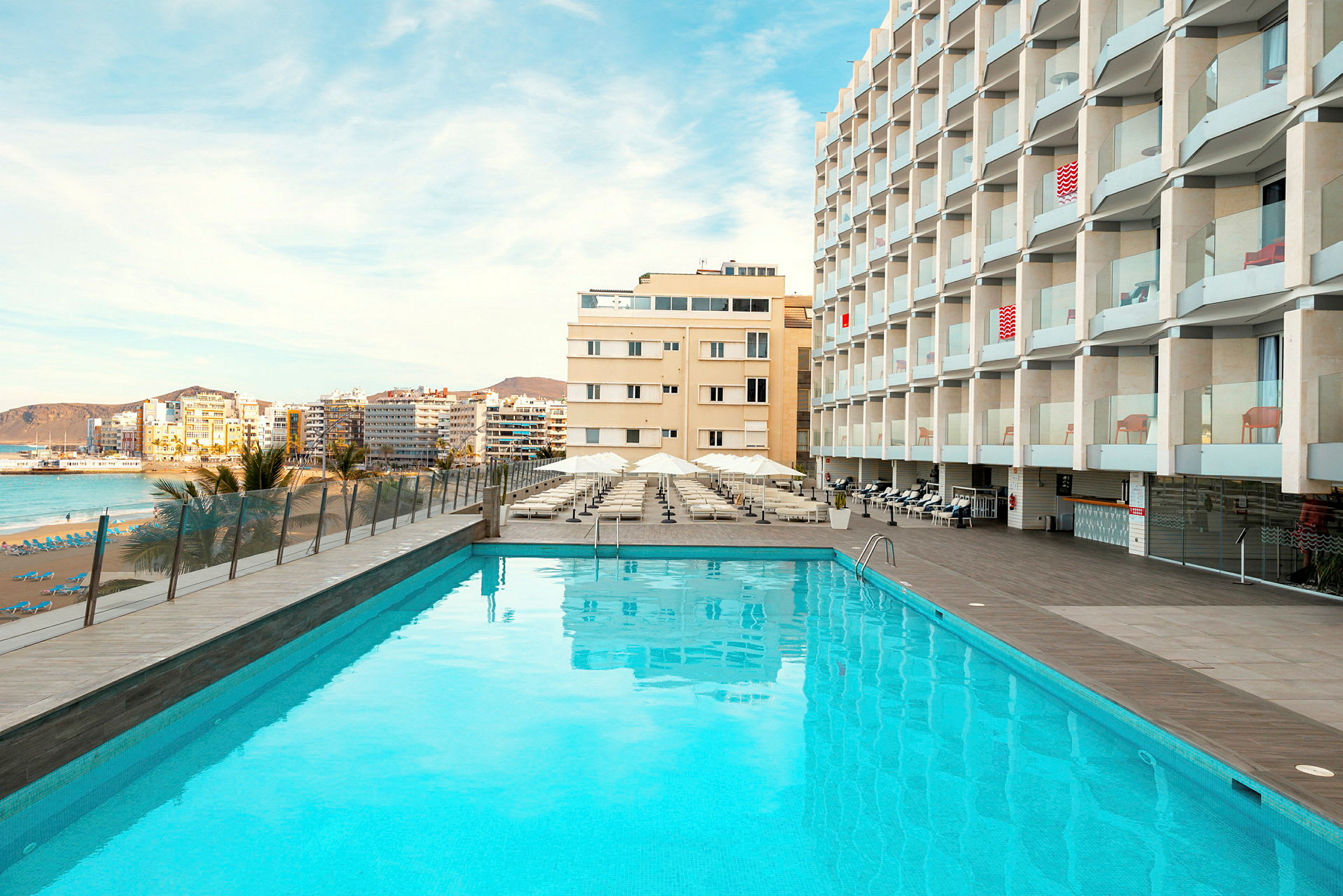 Utendørsbasseng foran hotellbygning, med strand og by i bakgrunnen. (AI-generert tekst)