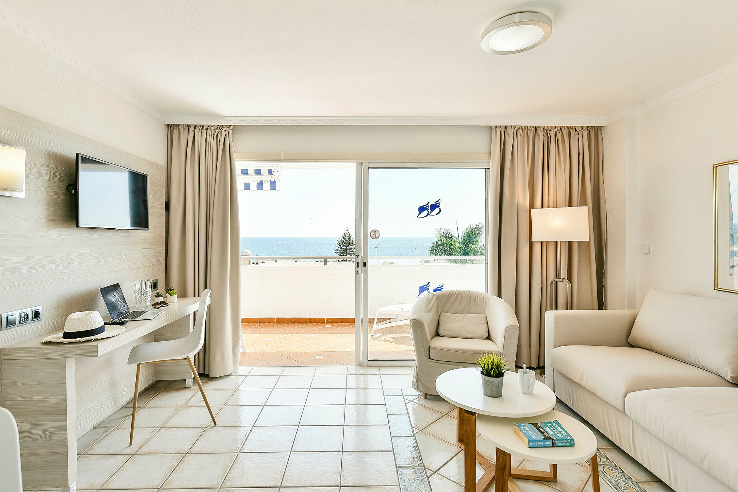 Lyst rom med sofa, lenestol, bord og utsikt mot havet gjennom glassdører. (AI-generert tekst)