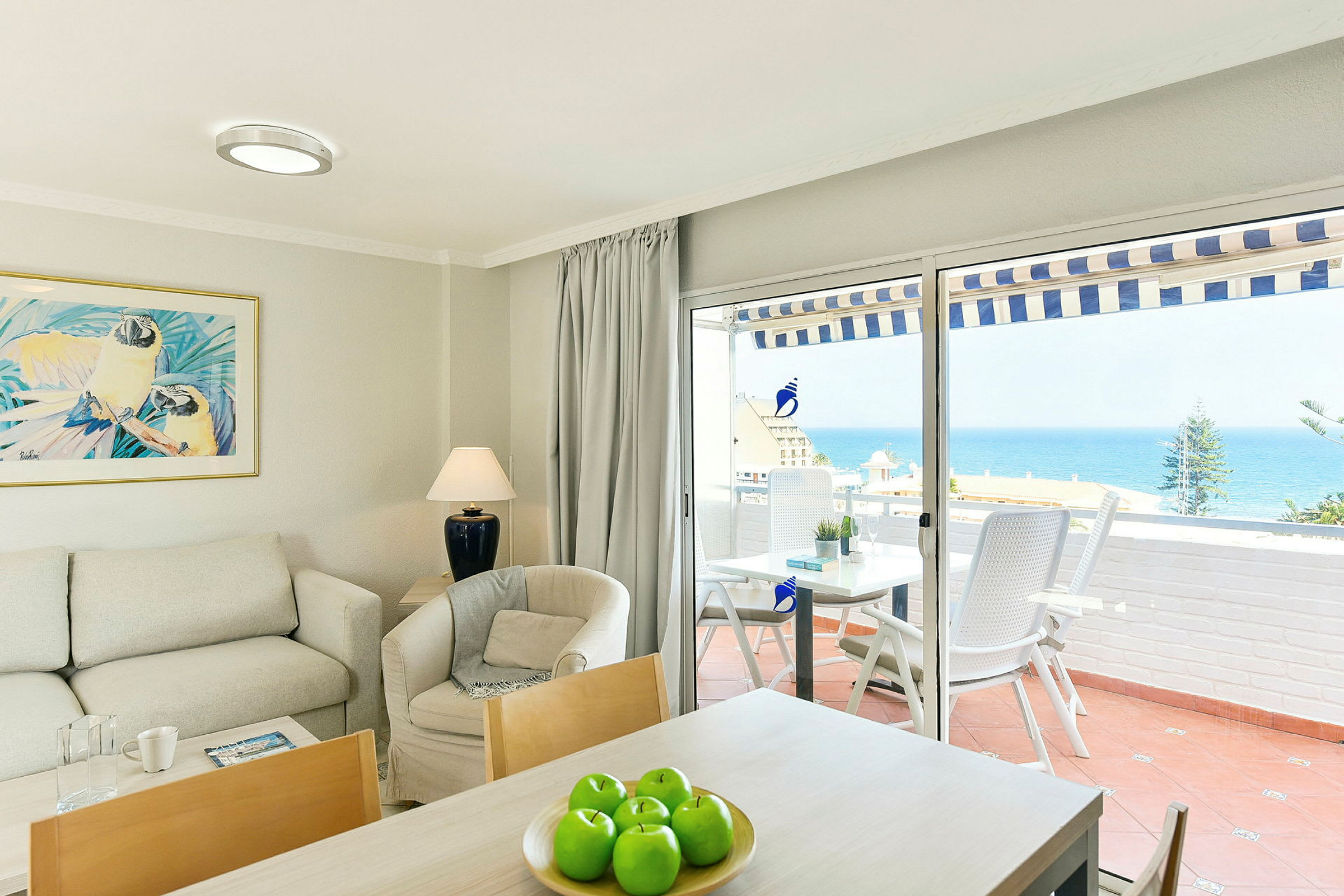 Interiør av en stue med sofa, lenestol og bord, og utsikt over havet gjennom glassdører til en terrasse. (AI-generert tekst)