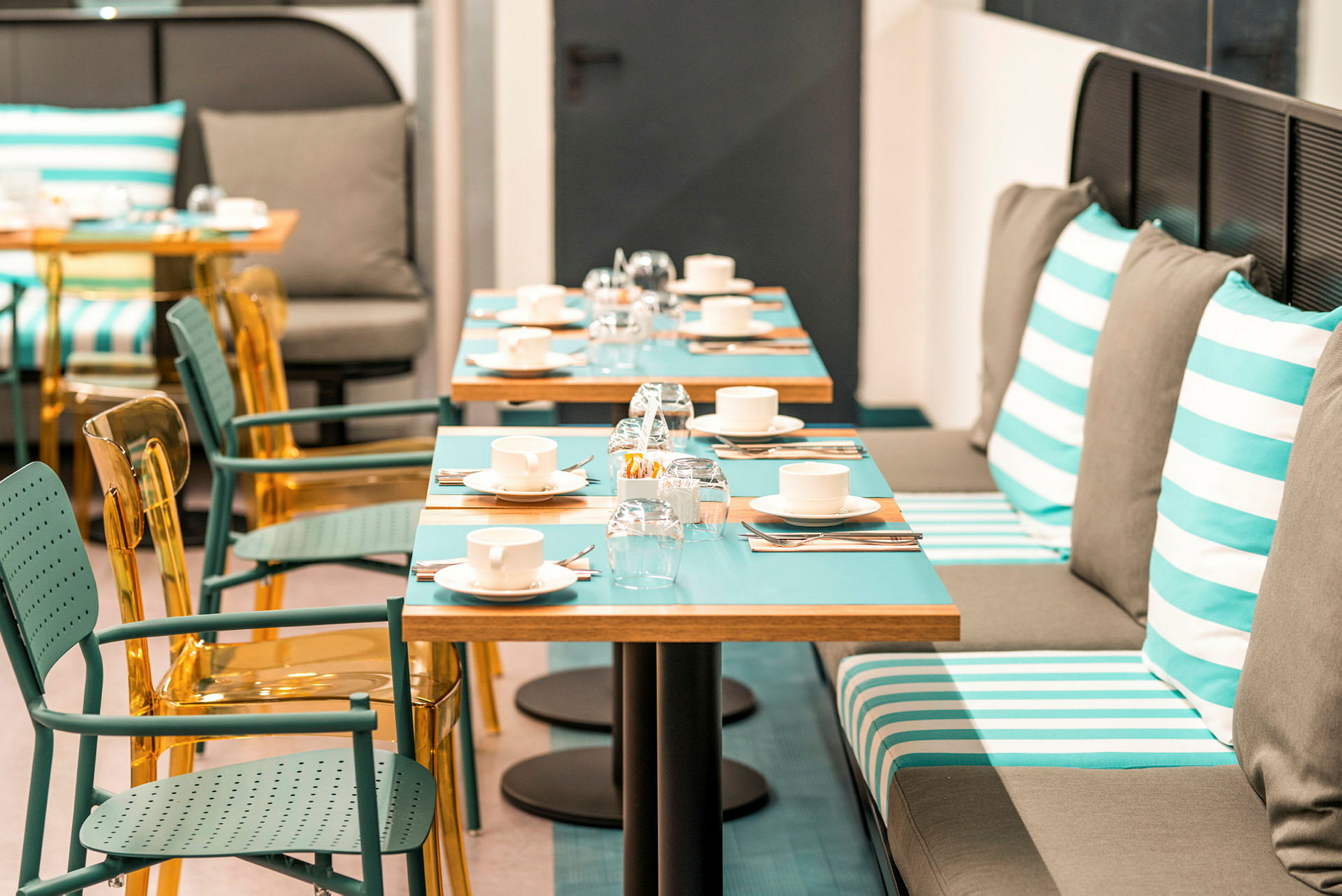 Bilde av en restaurant med bord dekket med kopper, glass og bestikk. Stoler og benkesitteplasser med stripete puter vises. (AI-generert tekst)
