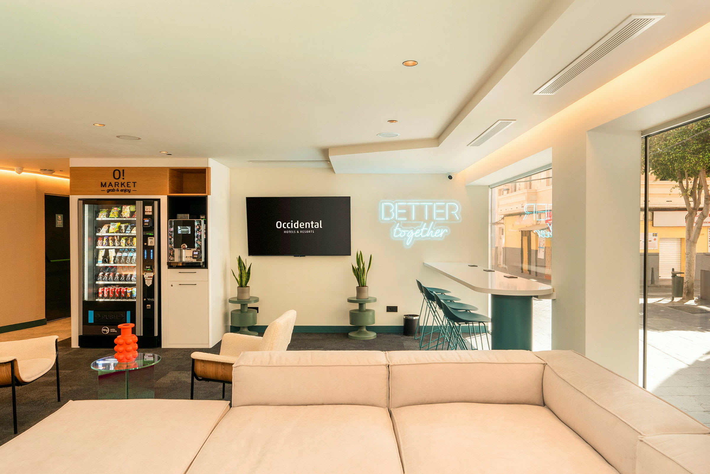 Et moderne og lyst loungeområde med sofa, stoler, bord og barstoler ved et høyt bord. Veggen har et skilt med teksten 'Better together' og en TV med logoen til Occidental Hotels & Resorts. (AI-generert tekst)