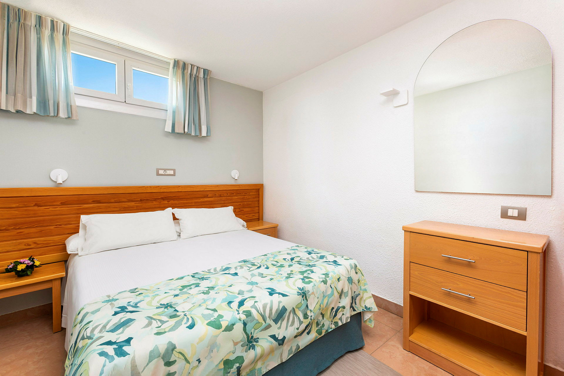 Et hotellrom med en dobbeltseng, trepanel som sengegavl, og et speil over en kommode. (AI-generert tekst)
