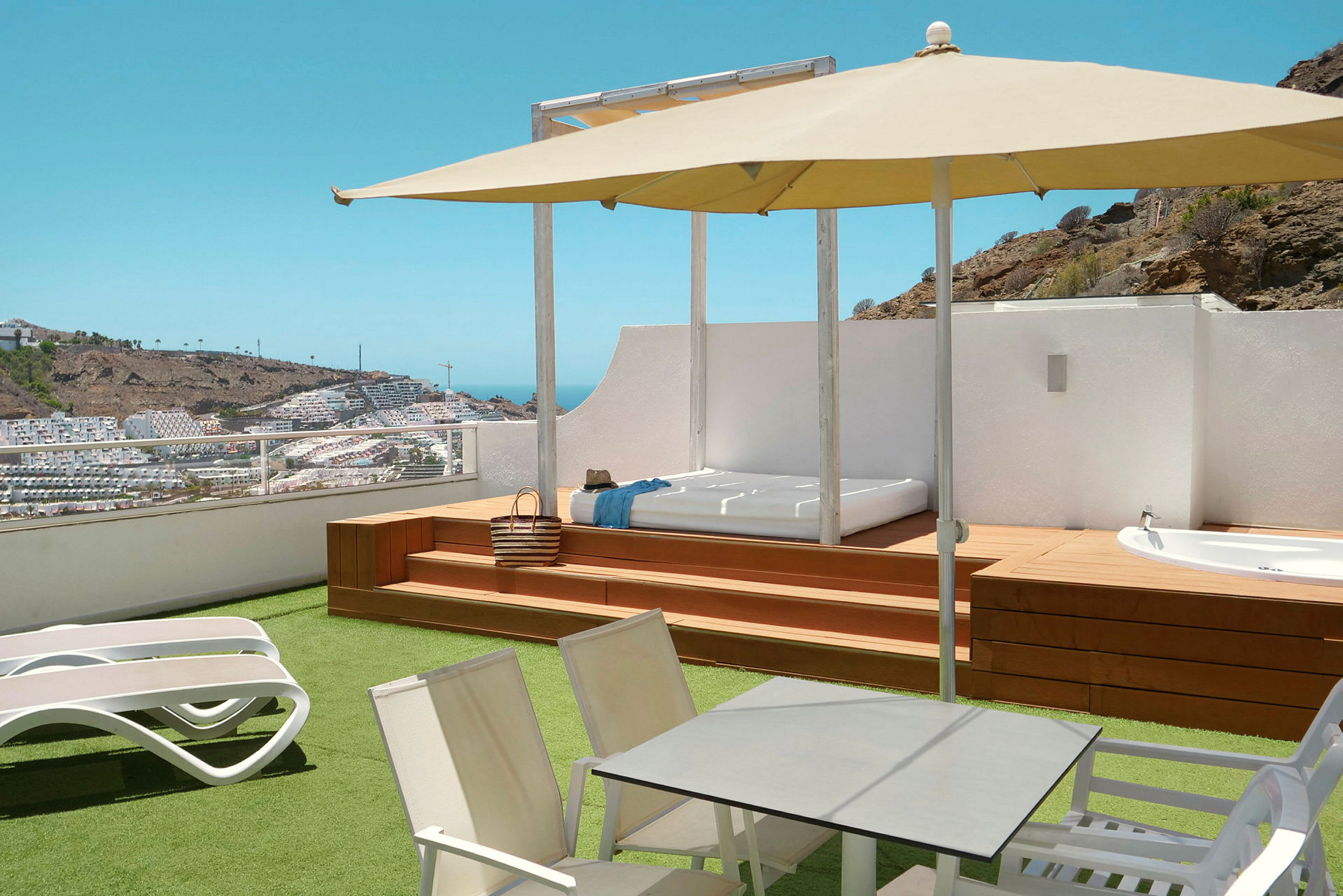 Terrasse med solsenger, bord, parasoll og utsikt over et kupert område med bygninger. (AI-generert tekst)