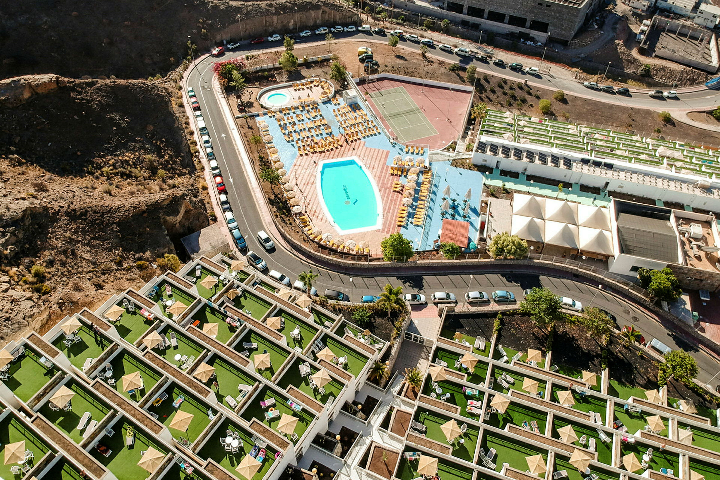 Luftfoto av hotellområde med basseng, solsenger og tennisbaner. (AI-generert tekst)
