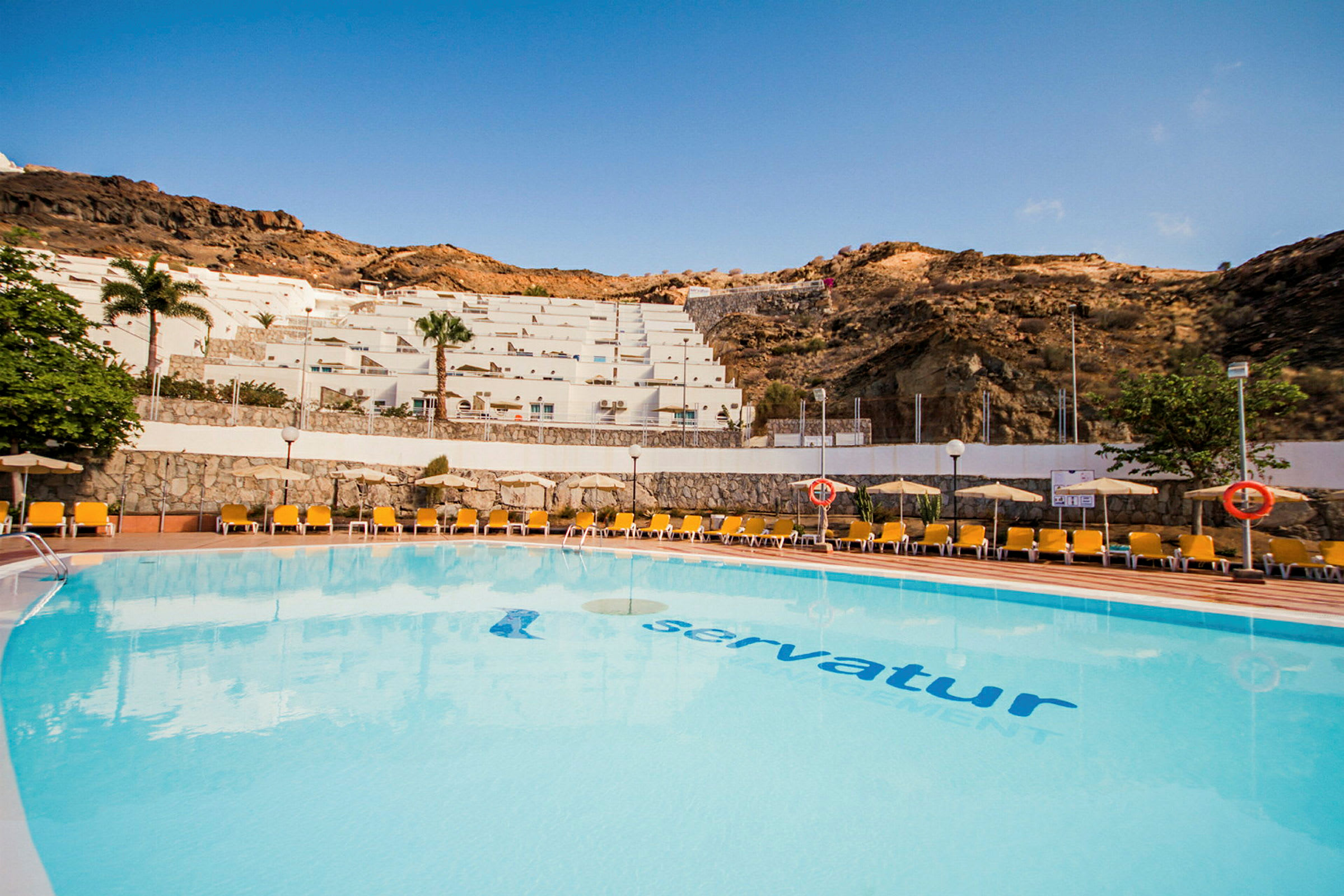 Utendørsbasseng ved et feriehotell med solsenger og utsikt mot en skråning med terrassebygninger. (AI-generert tekst)