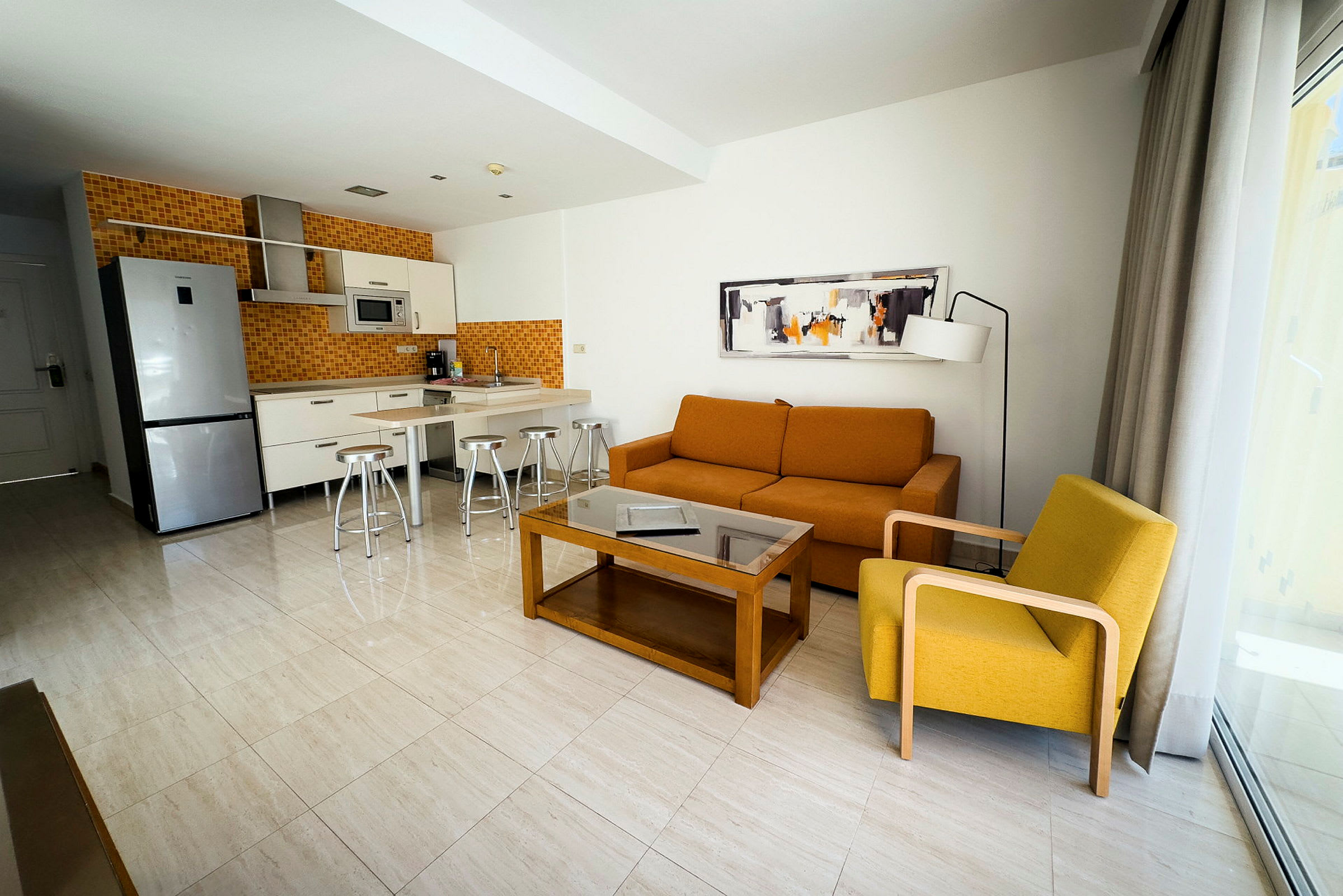 Moderne hotellrom med kjøkken og stue møbler inkludert oransje sofa og gul lenestol. (AI-generert tekst)