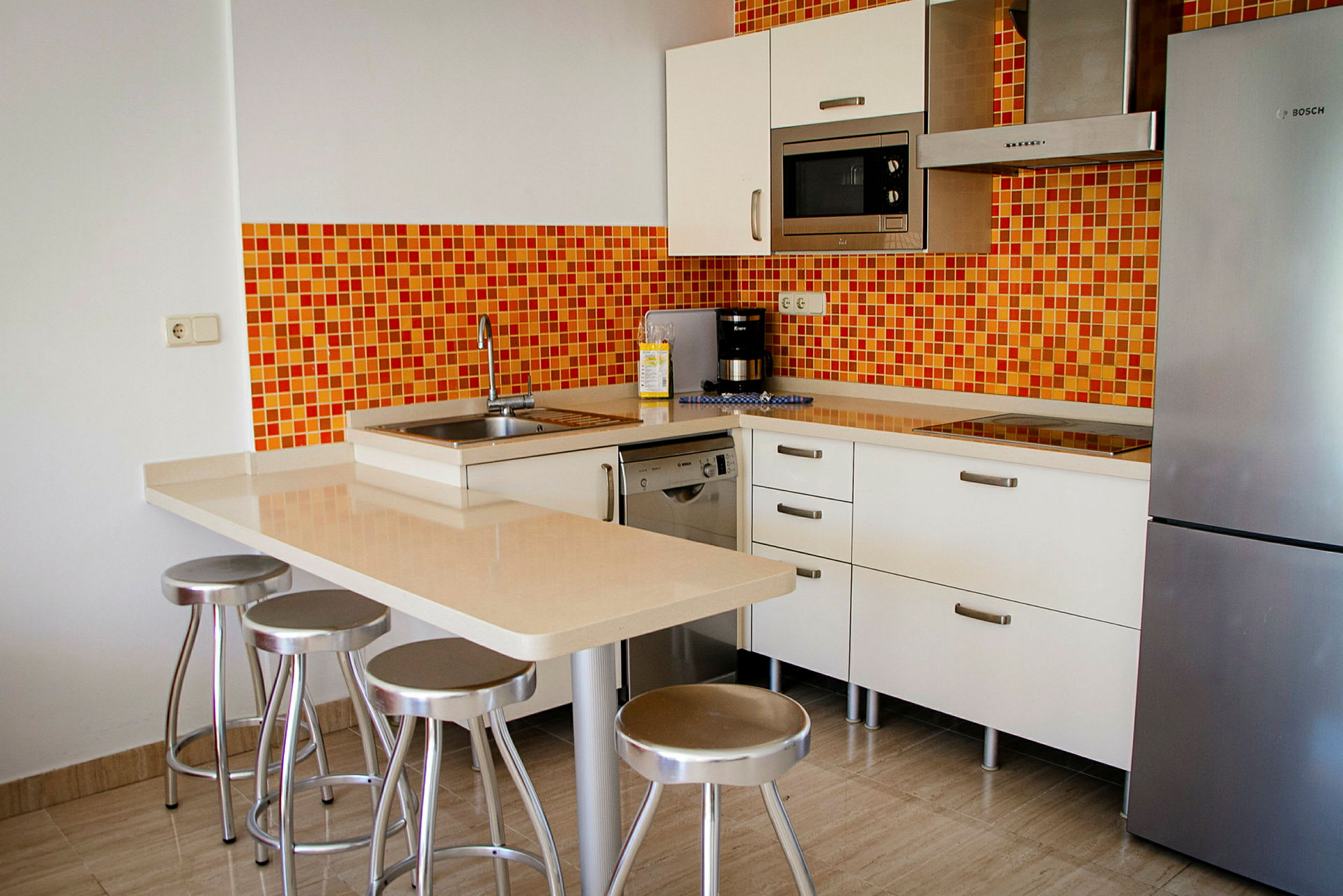 Moderne kjøkken med oransje mosaikkvegg, kjøleskap, mikrobølgeovn og en kjøkkenøy med barkrakker. (AI-generert tekst)