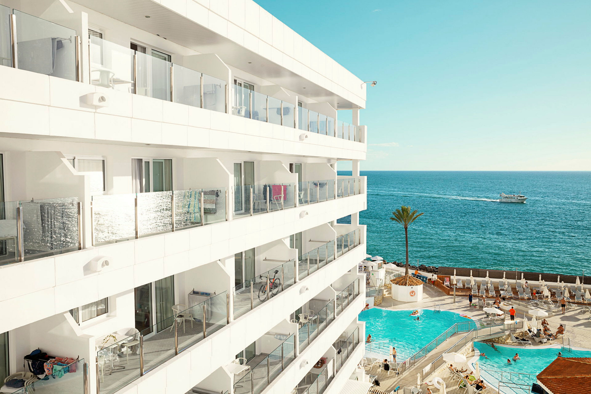 Utsikt over et moderne hotell med balkonger, et utendørs basseng og havet i bakgrunnen. (AI-generert tekst)