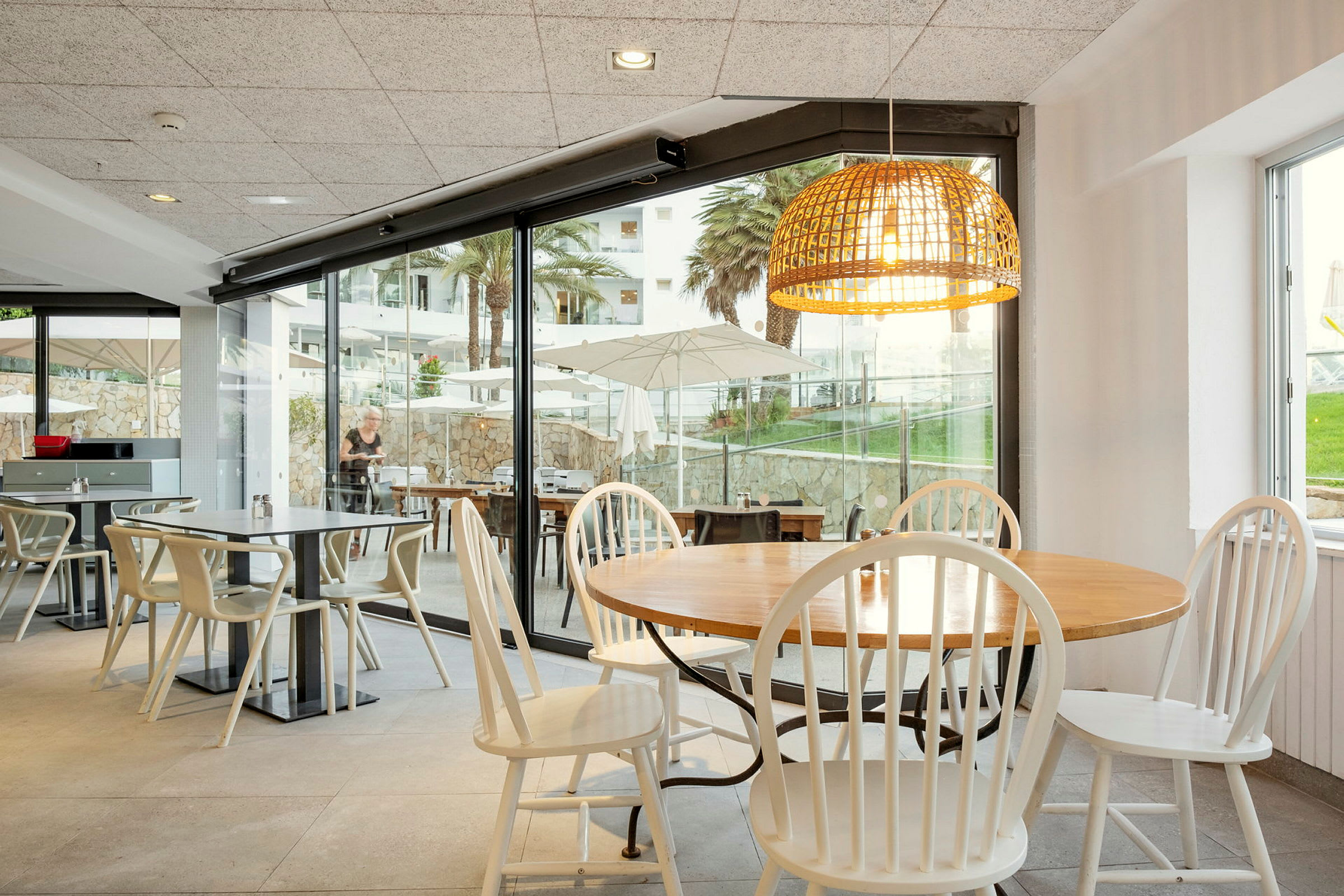 En restaurant med bord og stoler. Store glassvinduer viser utsikten med parasoller, grøntområder og palmer. (AI-generert tekst)