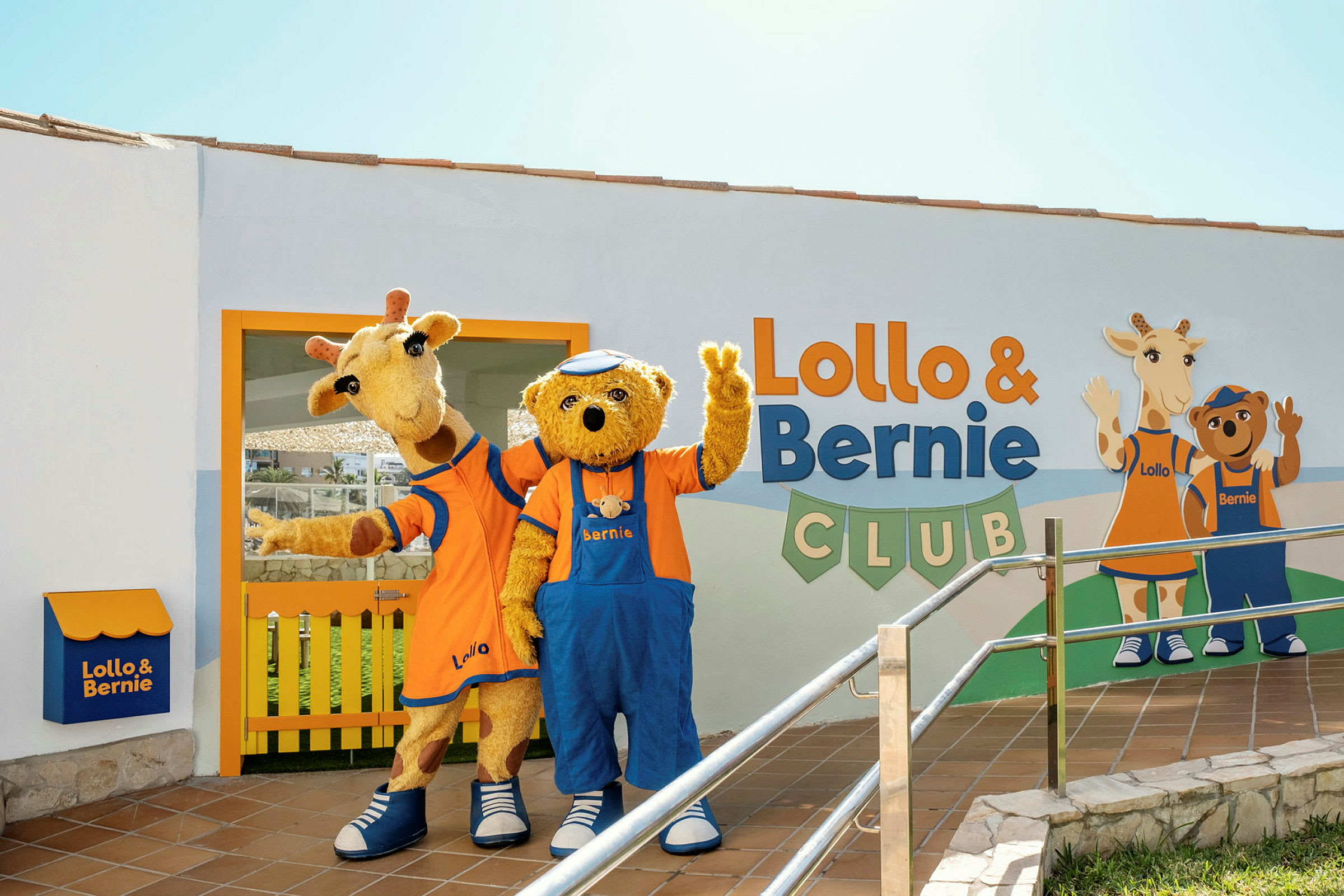 To maskoter, en giraff og en bjørn, står foran bygningen til Lollo & Bernie Club og vinker. (AI-generert tekst)