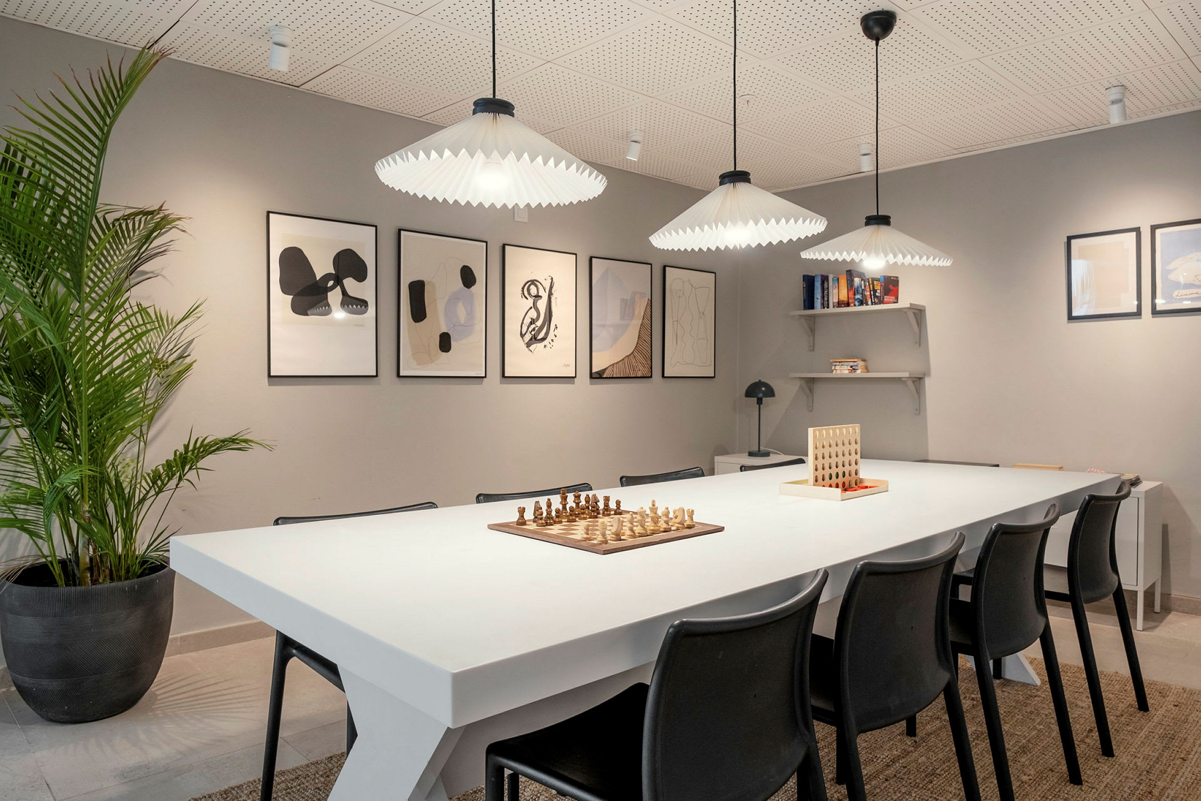 Bilde av et rom med et hvitt bord omgitt av svarte stoler. På bordet står et sjakkspill og et spill med brikker. Veggene er dekorert med innrammet abstrakt kunst, og en hylle inneholder bøker. (AI-generert tekst)