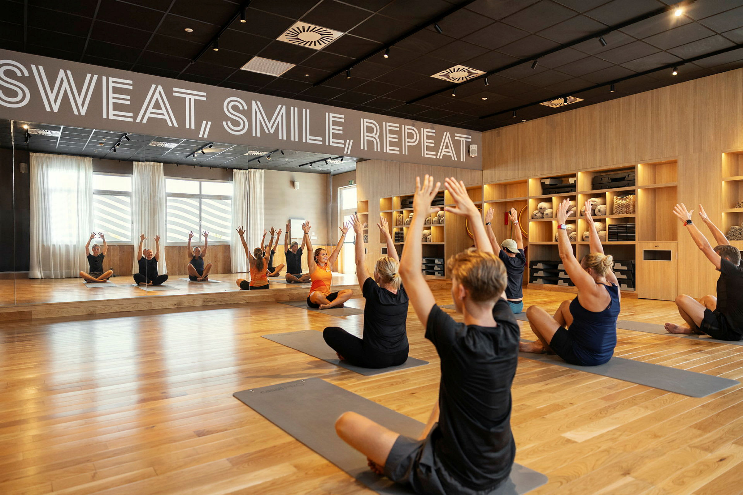 En treningssal med speilvegg der flere personer utfører stretchingøvelser på yogamatter. (AI-generert tekst)