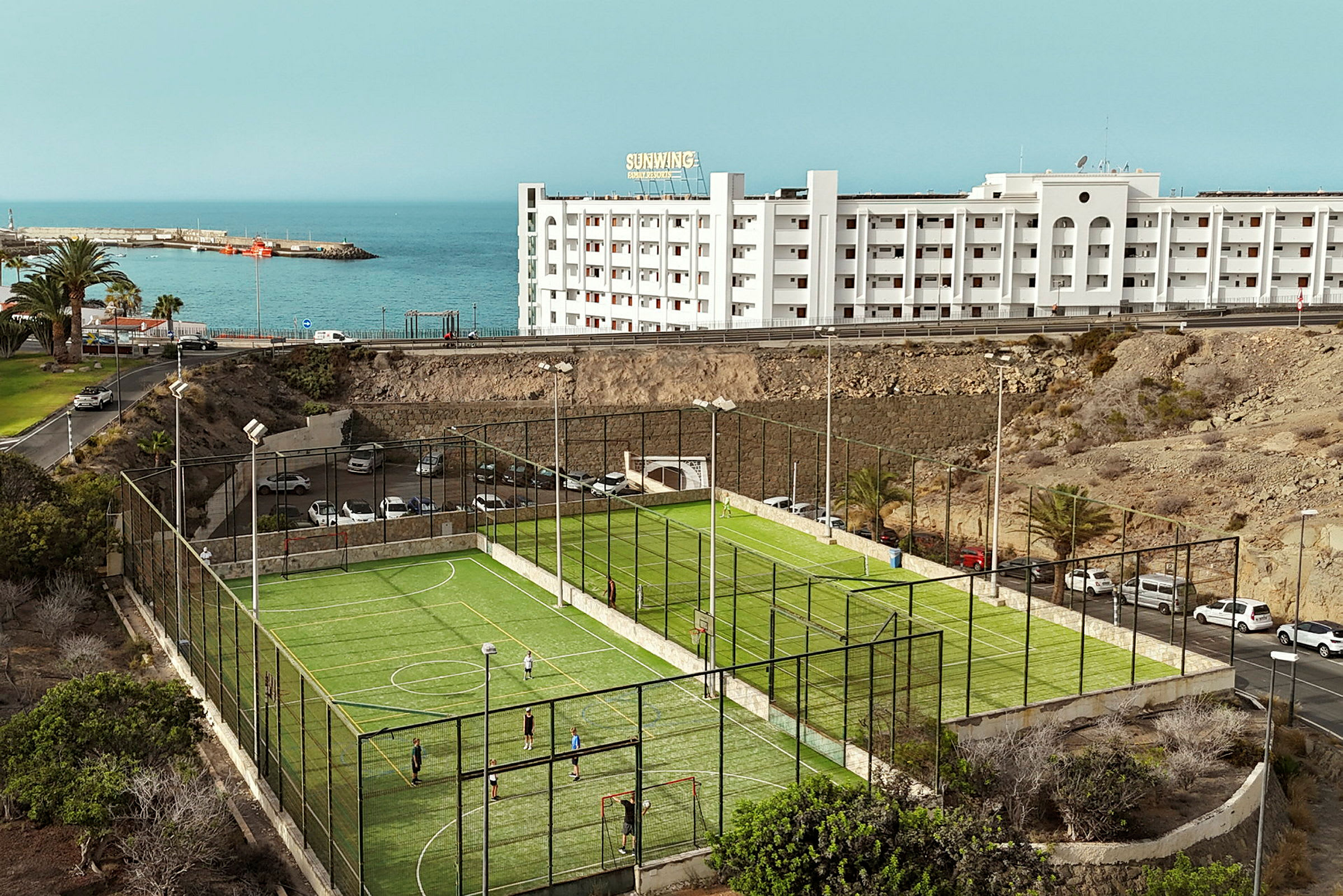 Fotballbane med grønt kunstgress ved kysten, bygning i bakgrunnen. (AI-generert tekst)