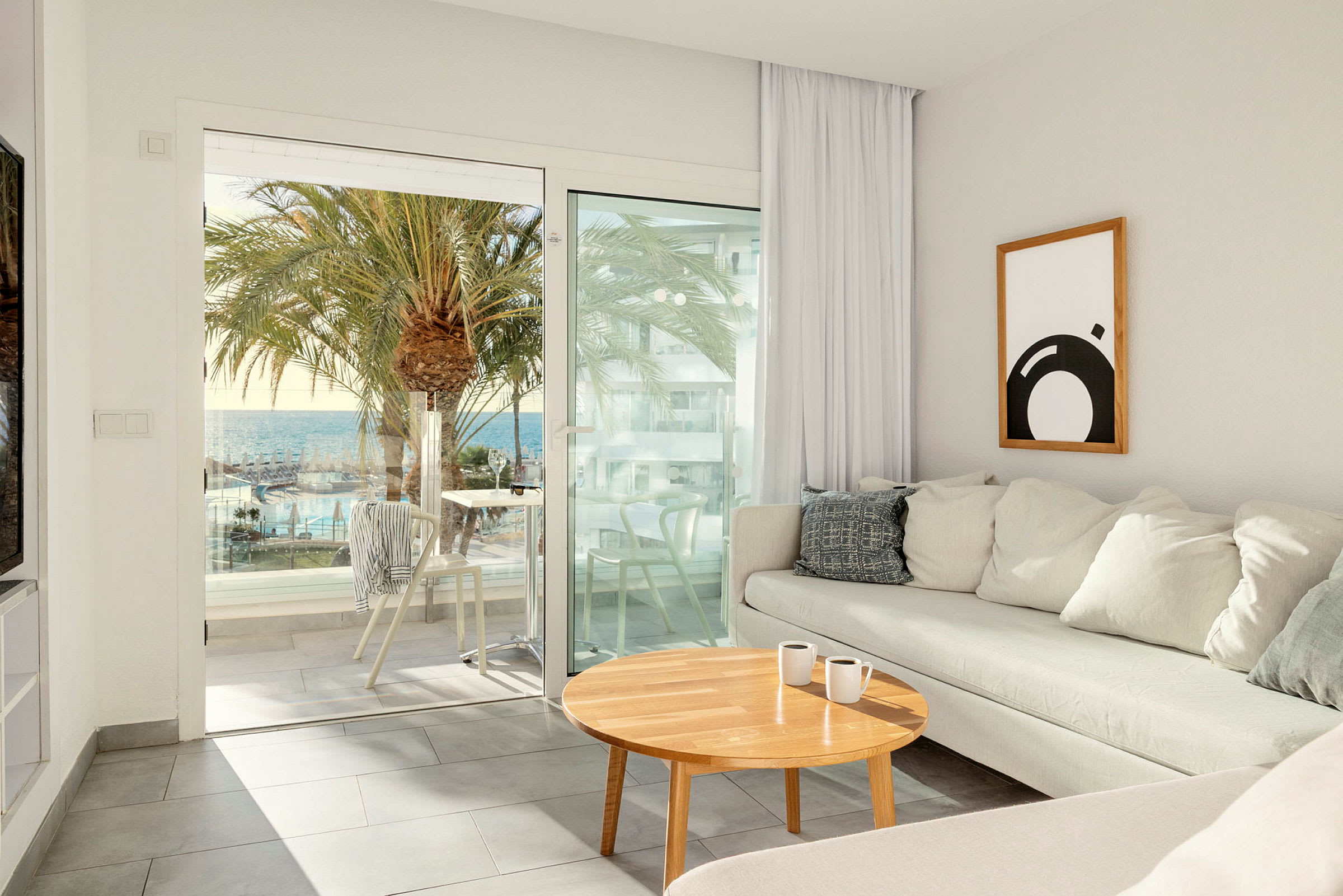 En lys stue med en beige sofa og et trebord har utsikt gjennom glassdører mot en terrasse med utsikt over palmer, et basseng og havet. (AI-generert tekst)