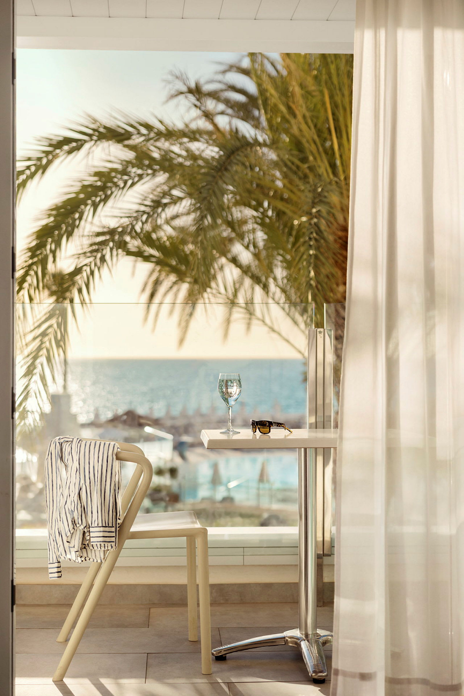 Et bord med et vinglass og et par solbriller står på en terrasse med utsikt over stranden og havet. En stol har en stripet skjorte hengende over ryggen og står ved siden av bordet. (AI-generert tekst)