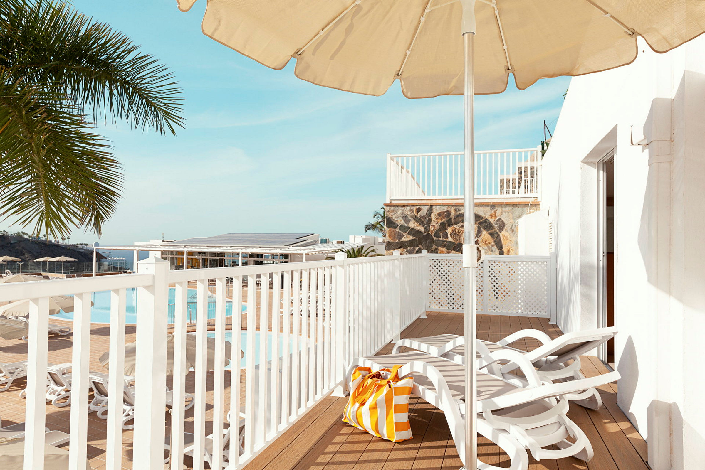 Utsikt over en terrasse med solsenger, parasoll og en strandveske. I bakgrunnen vises et basseng og palmer. (AI-generert tekst)