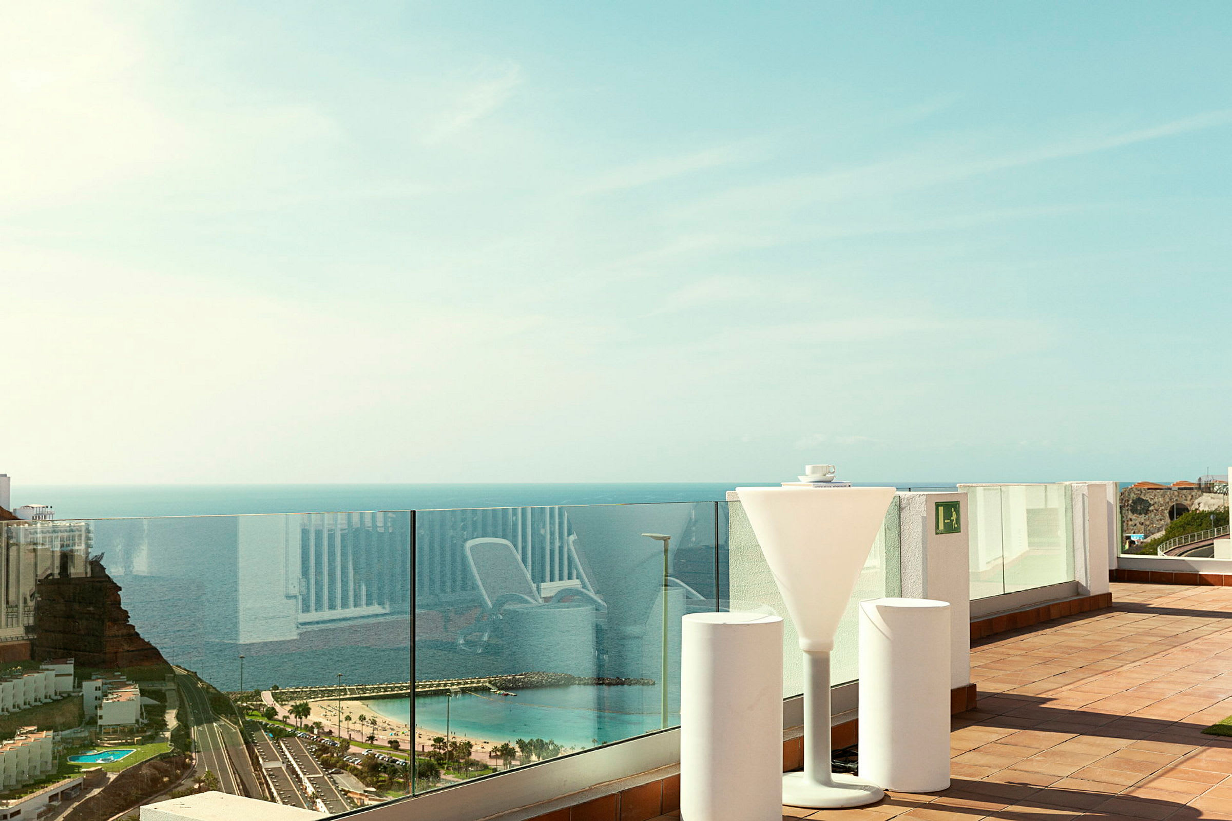 Utsikt fra en terrasse med glassrekkverk som viser havet og en strand. (AI-generert tekst)