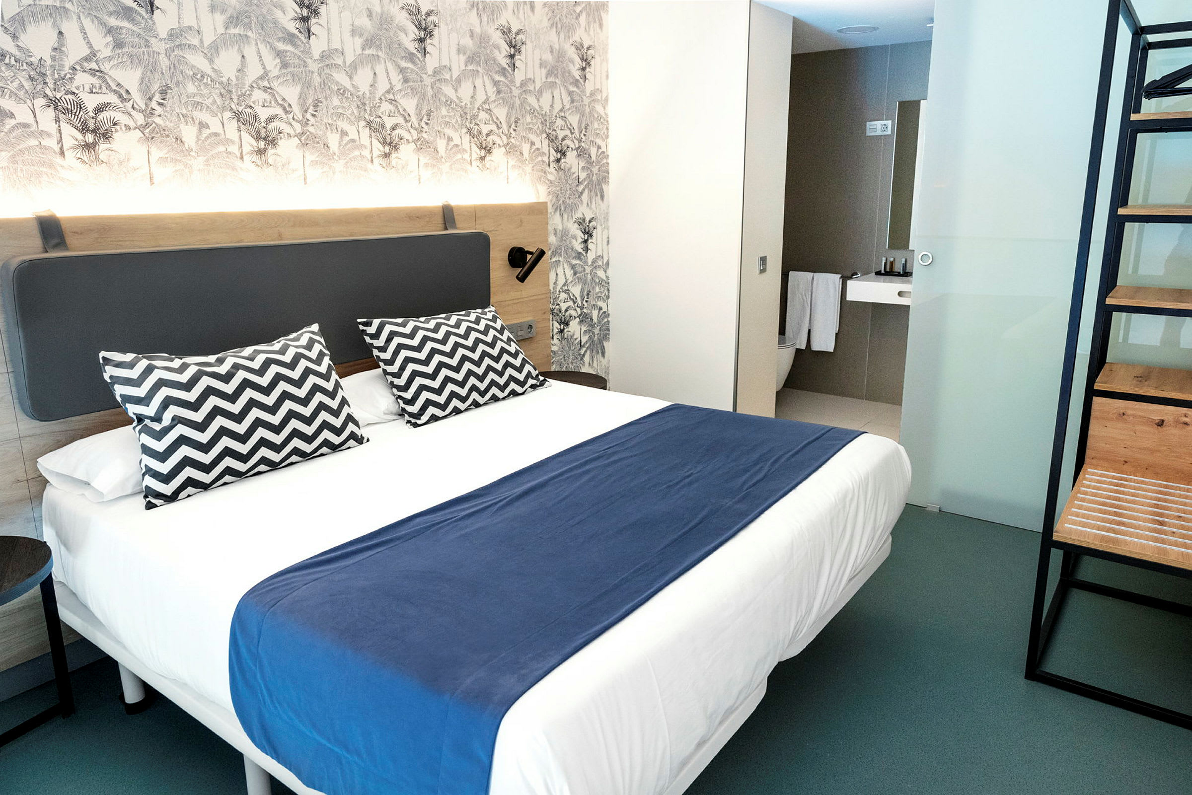 Et moderne hotellrom med dobbeltseng, blå sengeteppe og to mønstrede puter. (AI-generert tekst)