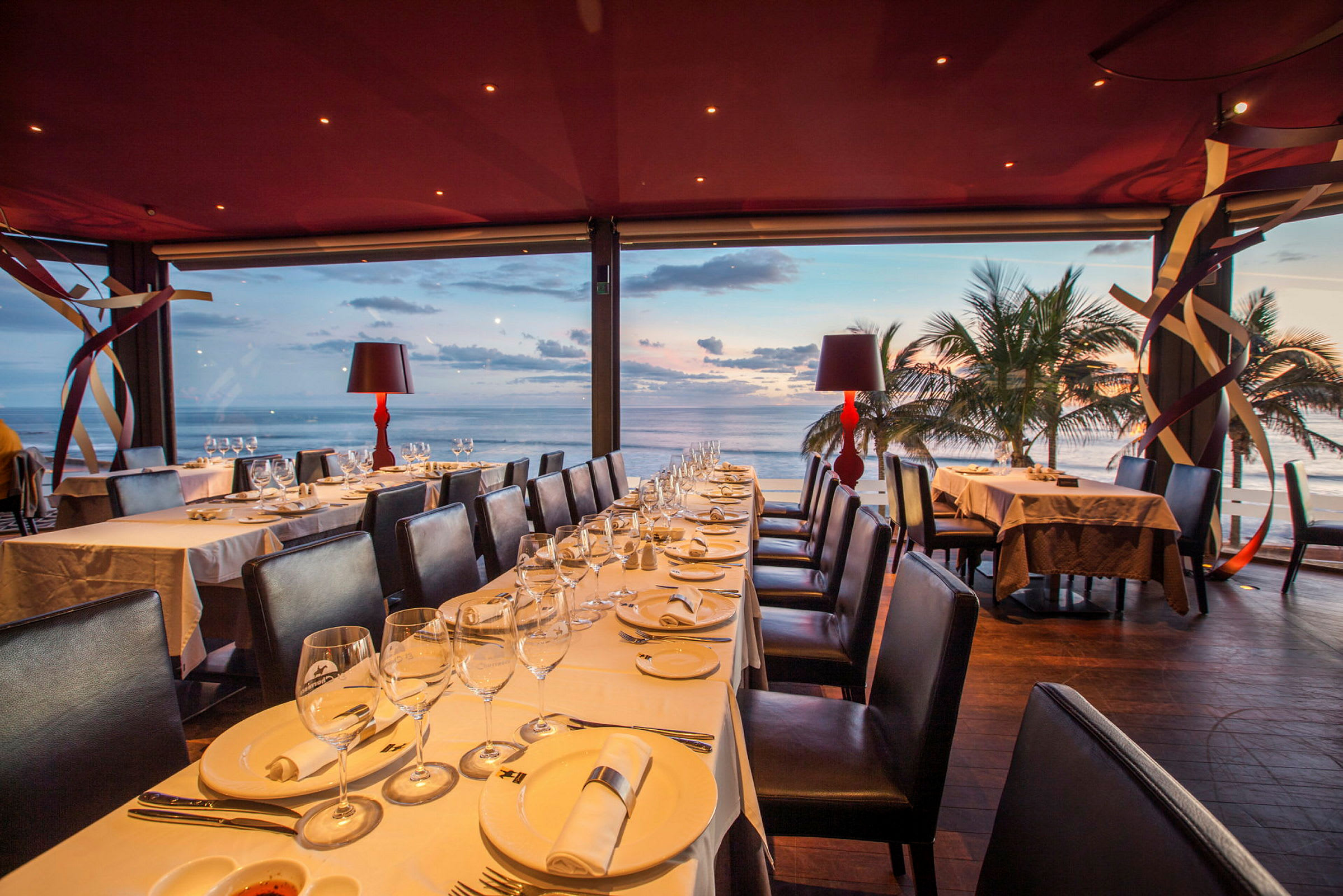 En restaurant med dekkede bord og utsikt over havet ved solnedgang. (AI-generert tekst)