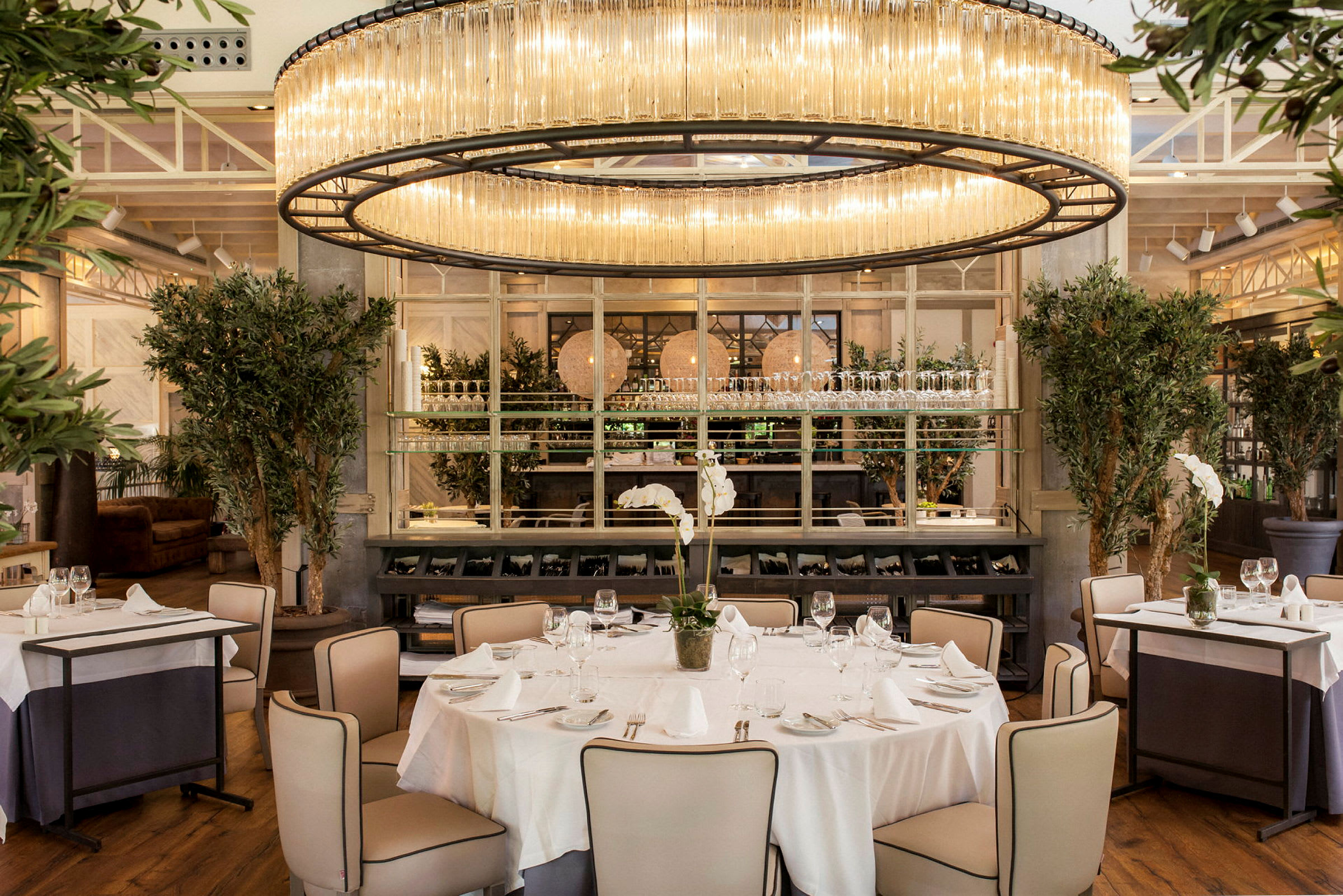 Elegant restaurantinteriør med en rund lampe i taket over et dekket bord omgitt av stoler. Rommet har også store planter og en moderne dekor. (AI-generert tekst)