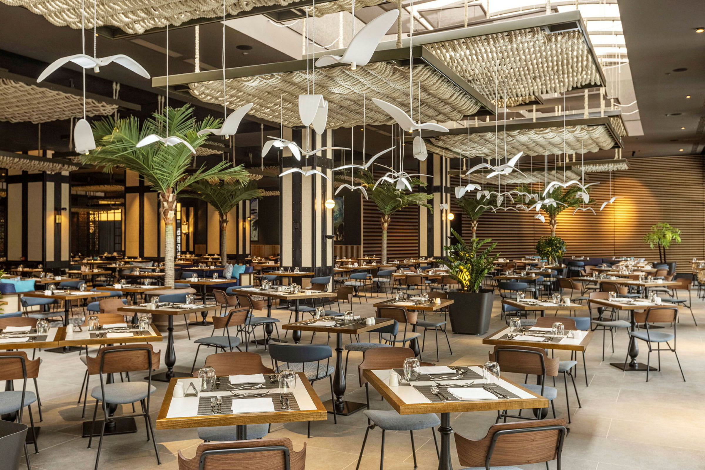 Restaurant med mange bord og stoler, dekorert med kunstneriske hengende elementer og planter. (AI-generert tekst)