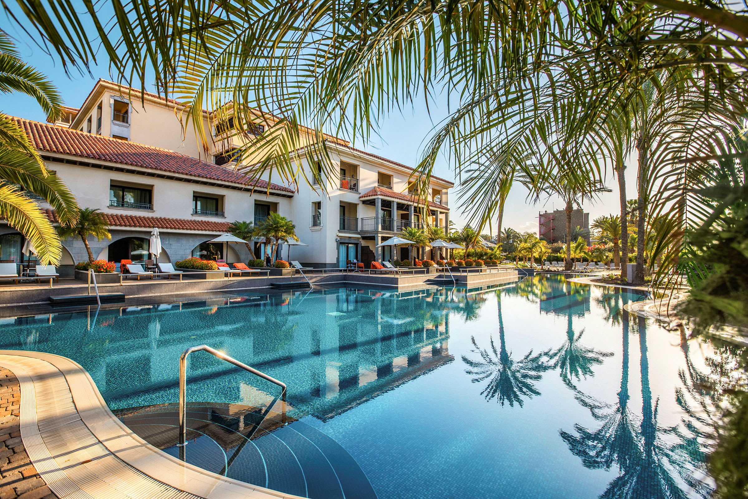 Et hotellbasseng omgitt av palmer og solsenger. (AI-generert tekst)
