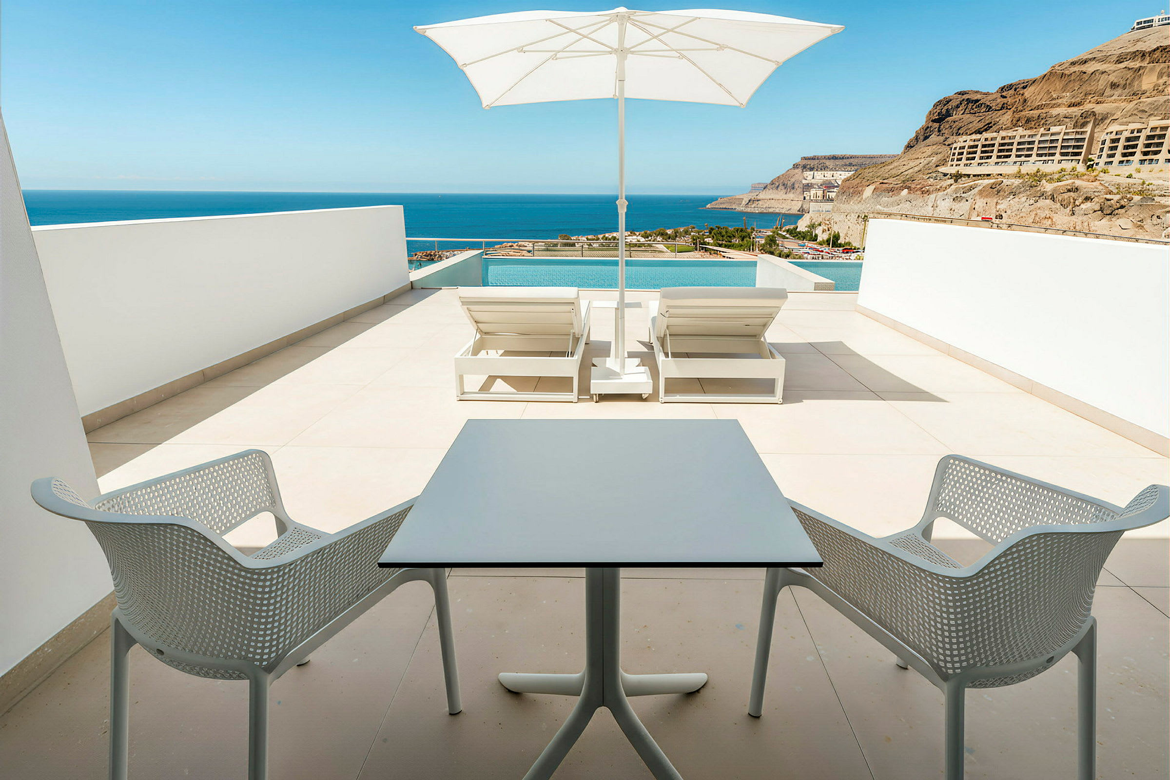En terrasse med to solsenger, parasoll og en liten sittegruppe med utsikt mot havet. (AI-generert tekst)