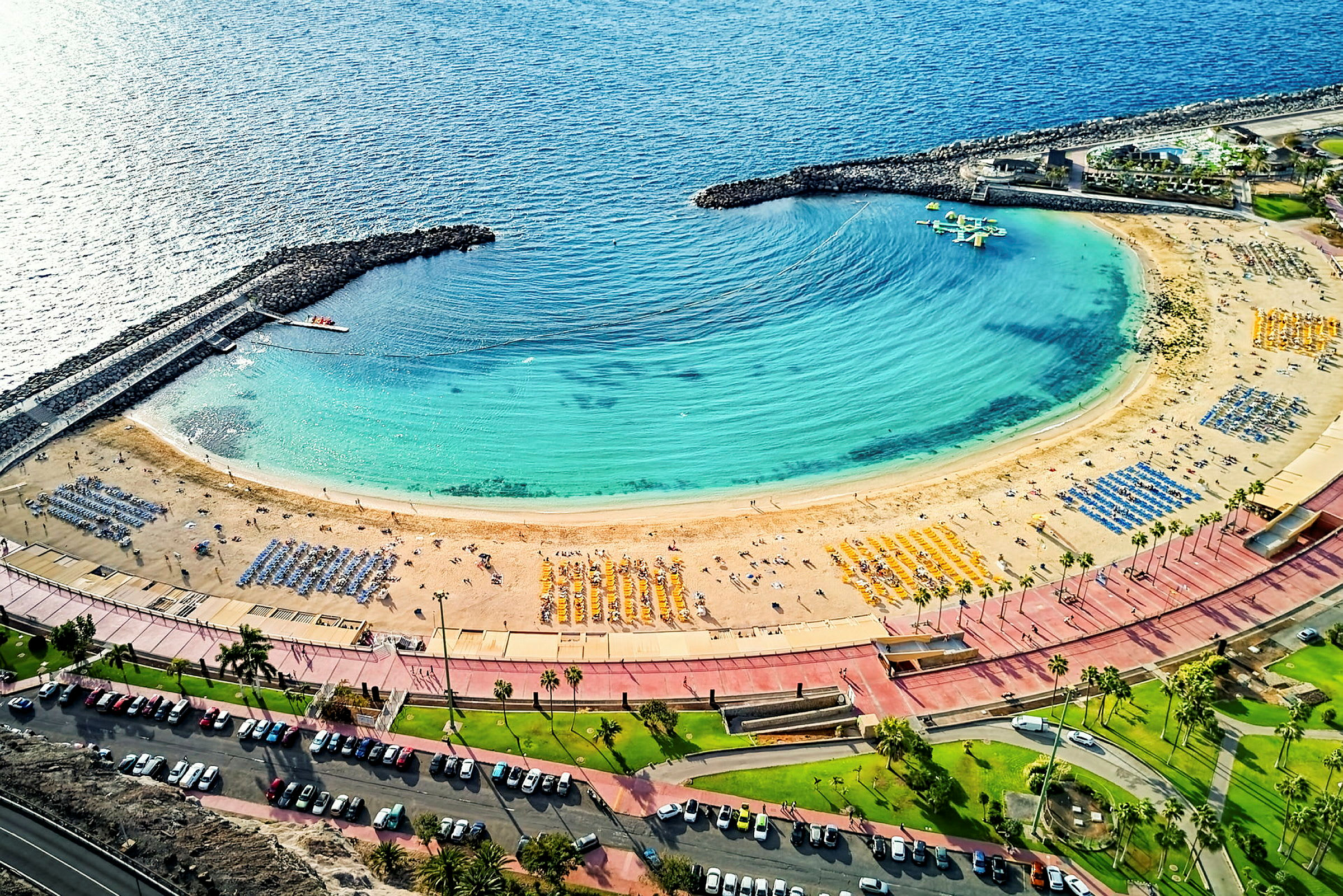 Fugleperspektiv av en velordnet sandstrand med parasoller, omgitt av klart blått vann og infrastruktur langs stranden. (AI-generert tekst)